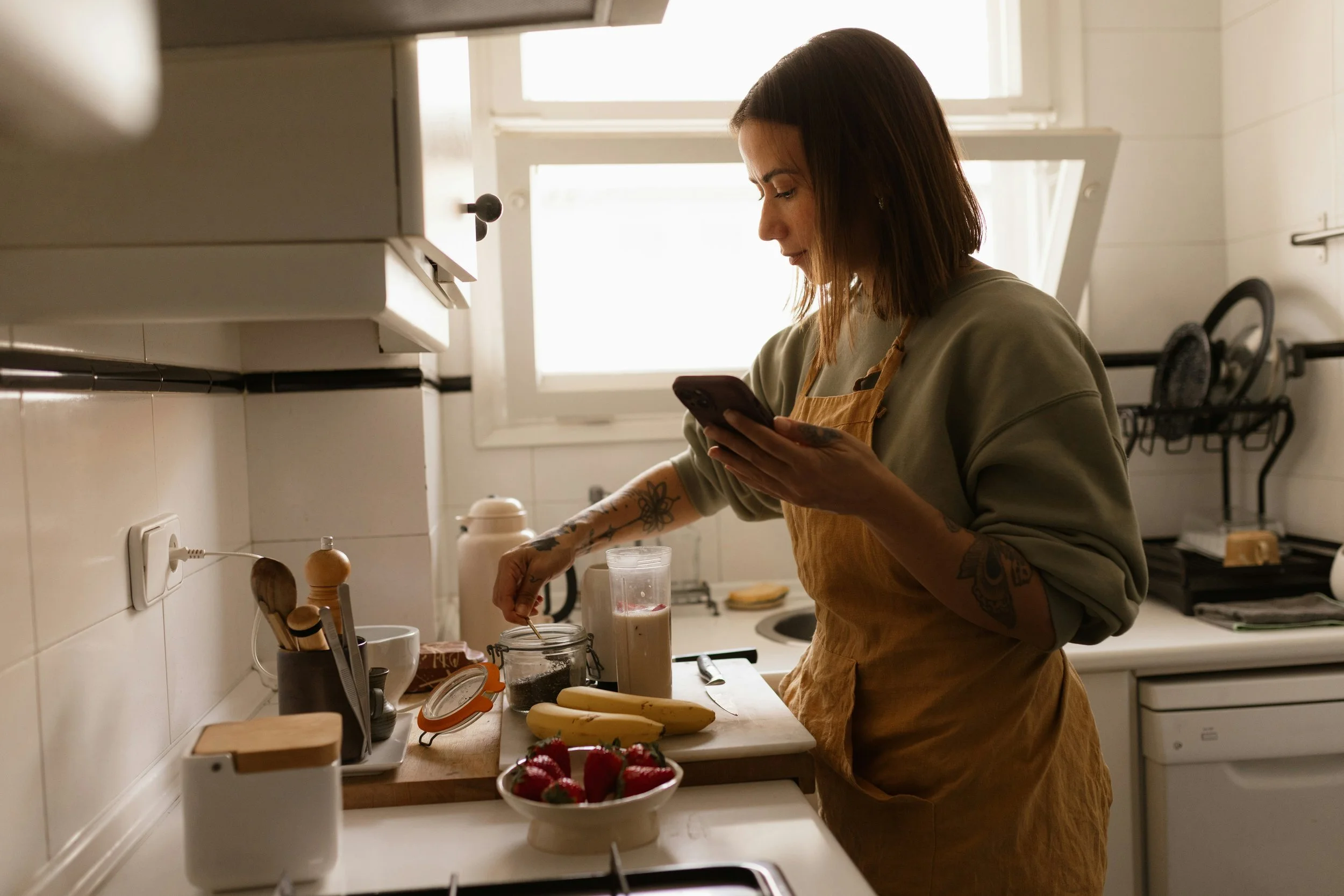 A woman in an apron making breakfast in a sunny kitchen while scrolling her phone Image title: Scrolling the feed while making breakfast