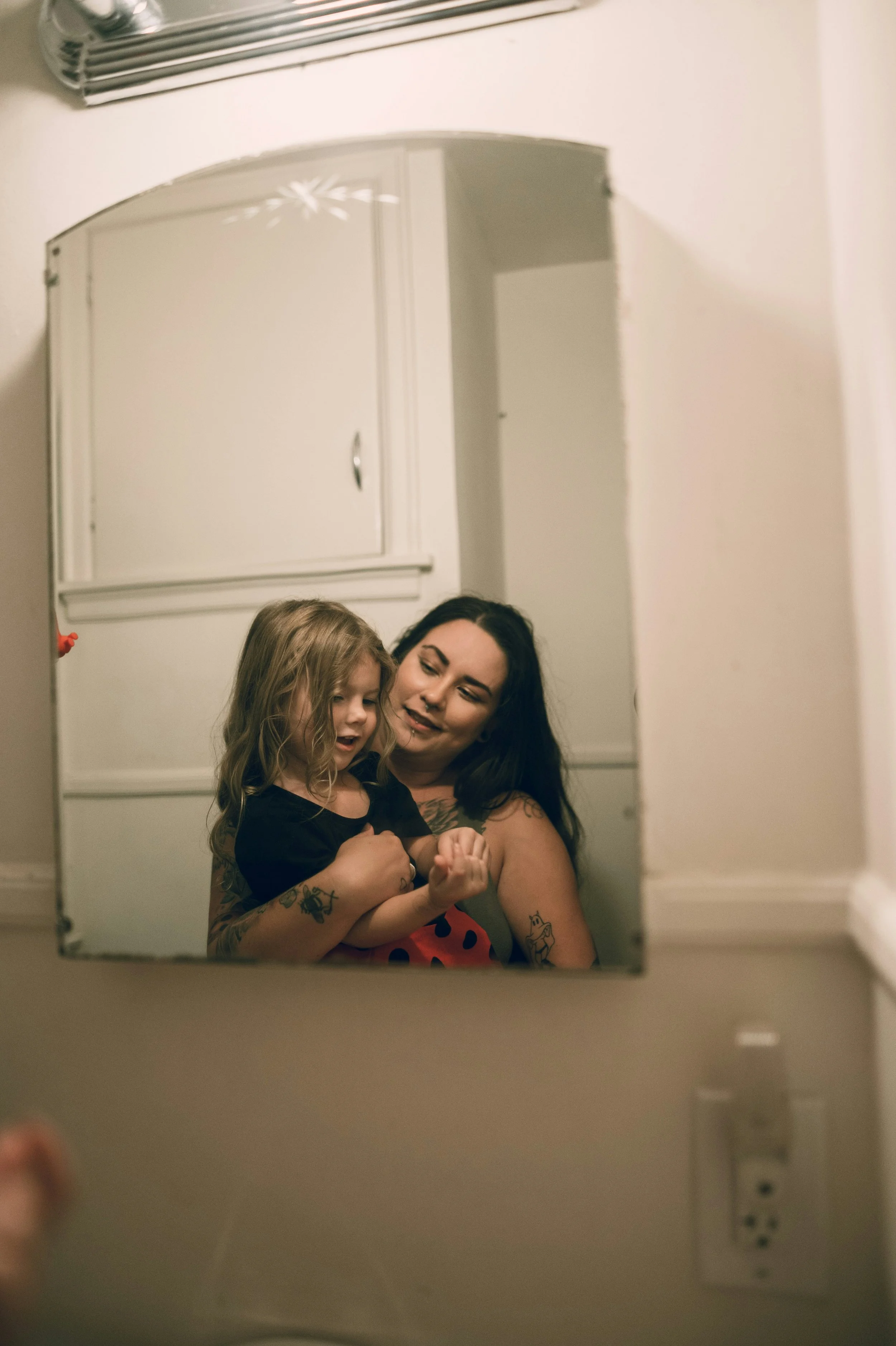 A tattooed mother holds her young daughter in her arms, their reflection captured in a vintage arched bathroom mirror under warm overhead light.