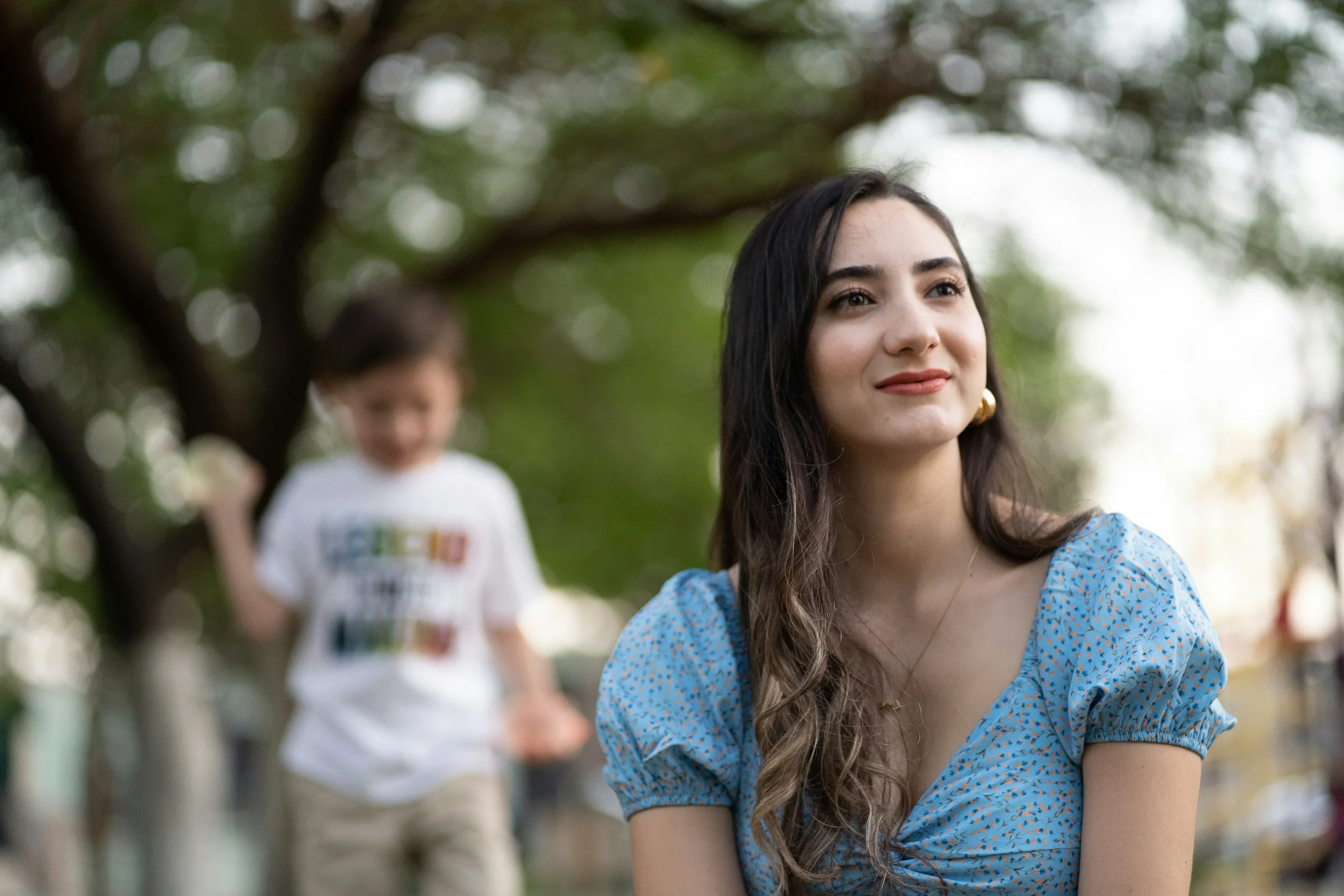 A mother smiling softly in a park with her child playing in the background