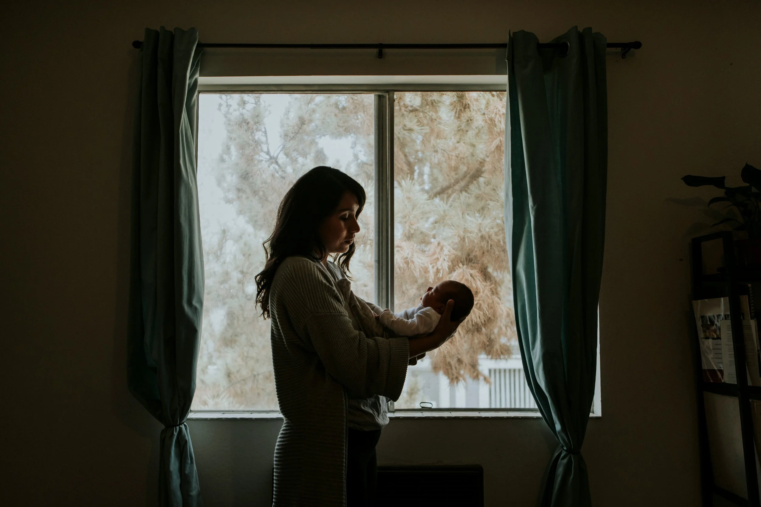 Mother standing at a window in a dim room, holding her newborn