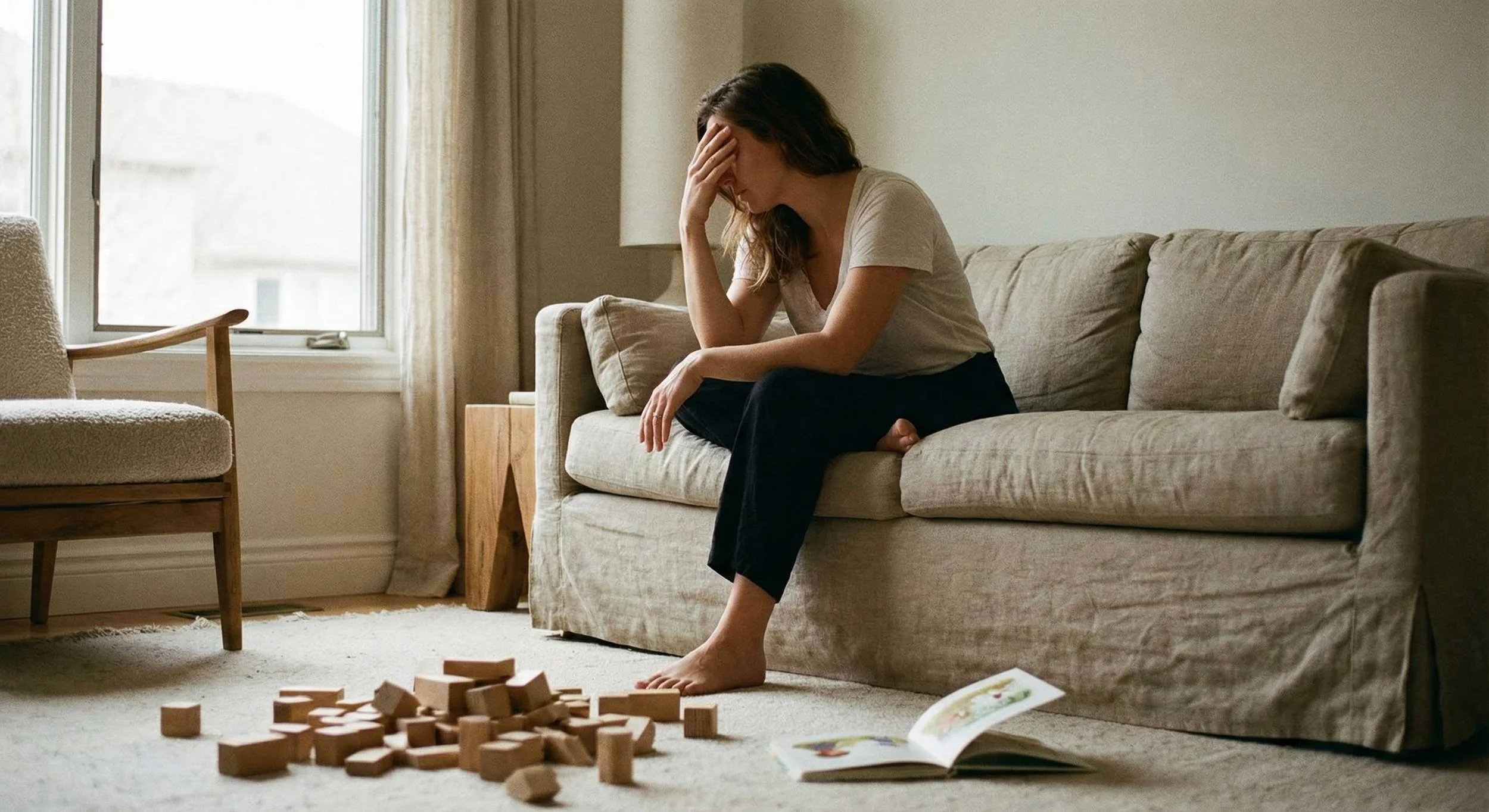 A mother sits on a beige couch covering her eyes with one hand in a posture of exhaustion. Scattered wooden building blocks and an open children's book are on the rug in the foreground.