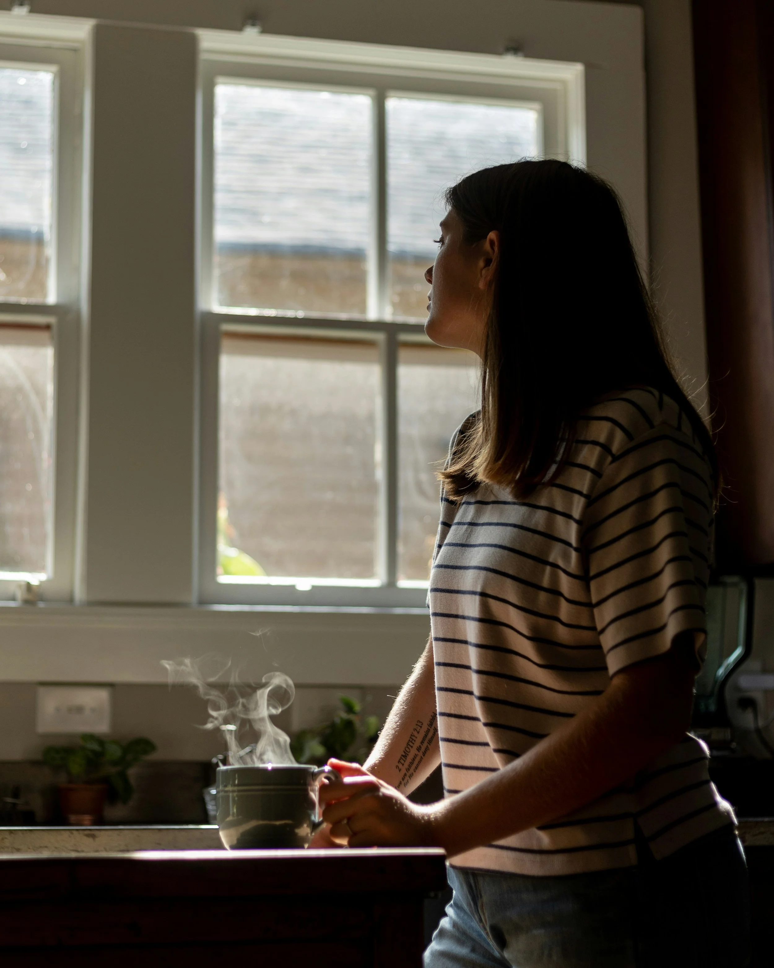 A woman holding a steaming mug by a sunlit kitchen window, looking out in a moment of stillness