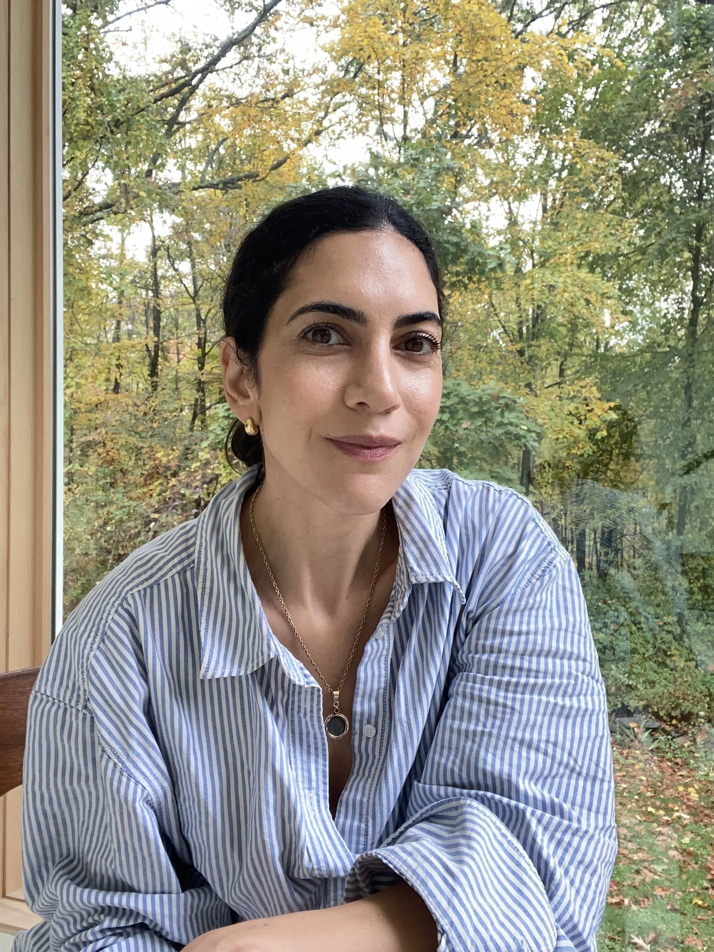 A woman with dark hair and light skin, wearing a blue and white striped shirt and gold jewelry, sitting inside near a window with an autumnal forest view outside.
