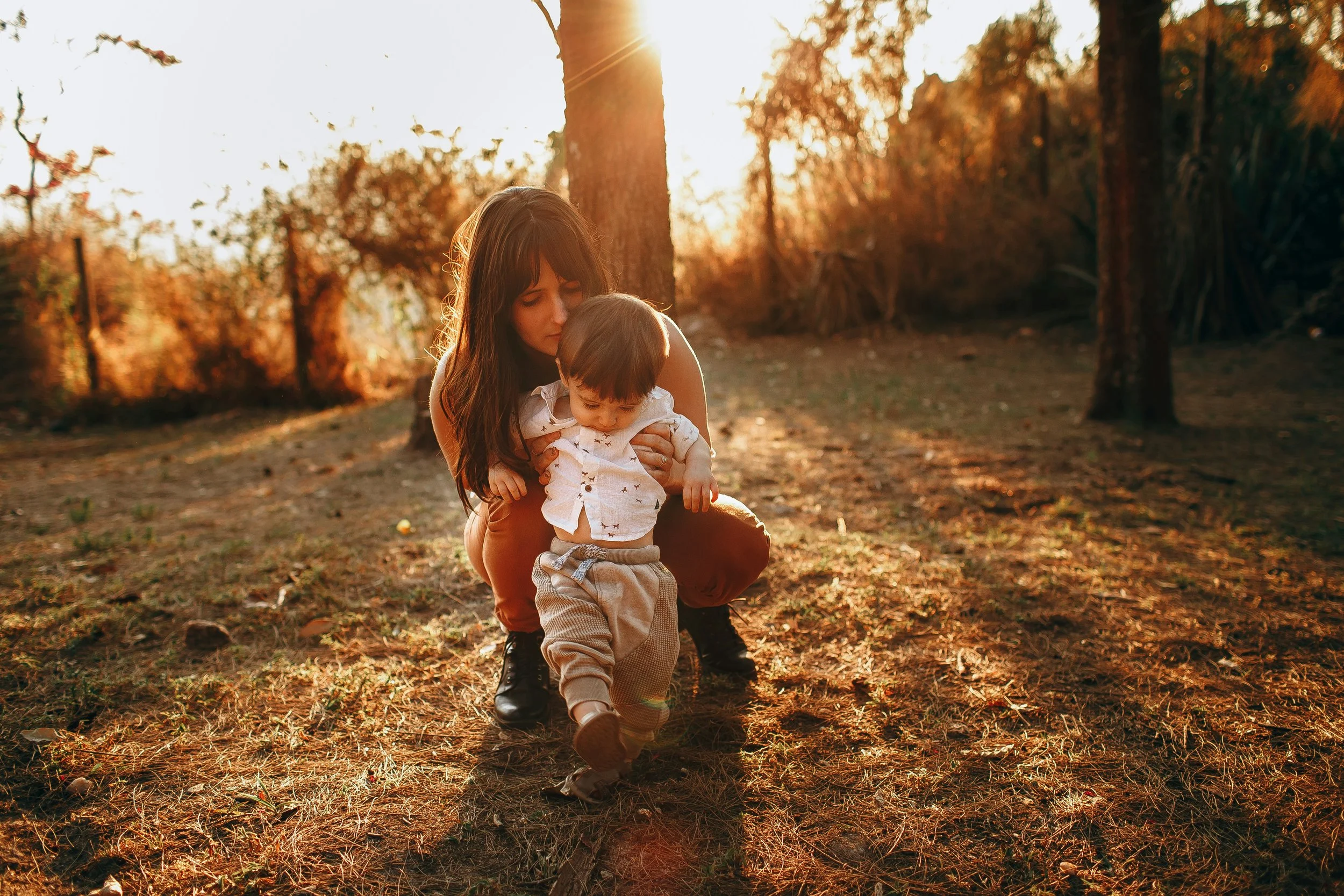Mother crouching behind her toddler outdoors in warm autumn light, holding him close and kissing the top of his head.