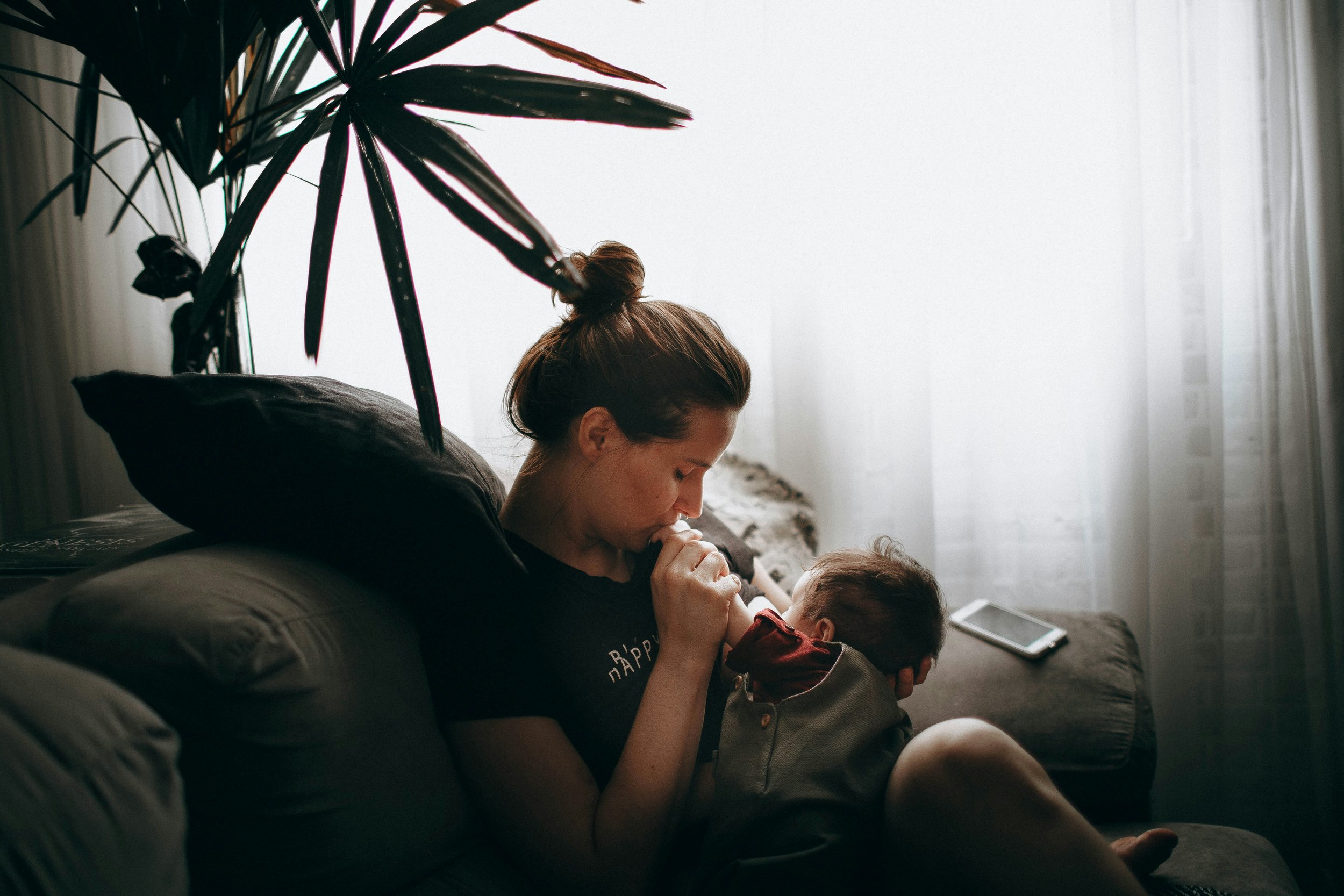 A mother sitting on a sofa in soft window light, kissing her baby's hand.