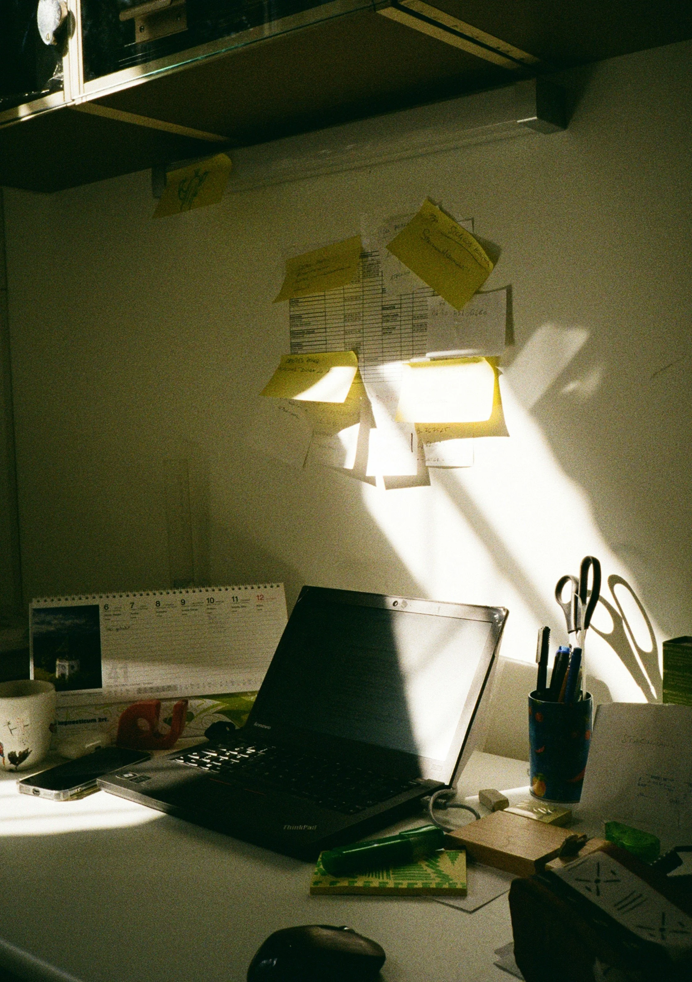 A cluttered desk bathed in morning sunlight with an open laptop, sticky notes on the wall, and a calendar, representing the invisible mental load carried by high-achieving mothers during matrescence.