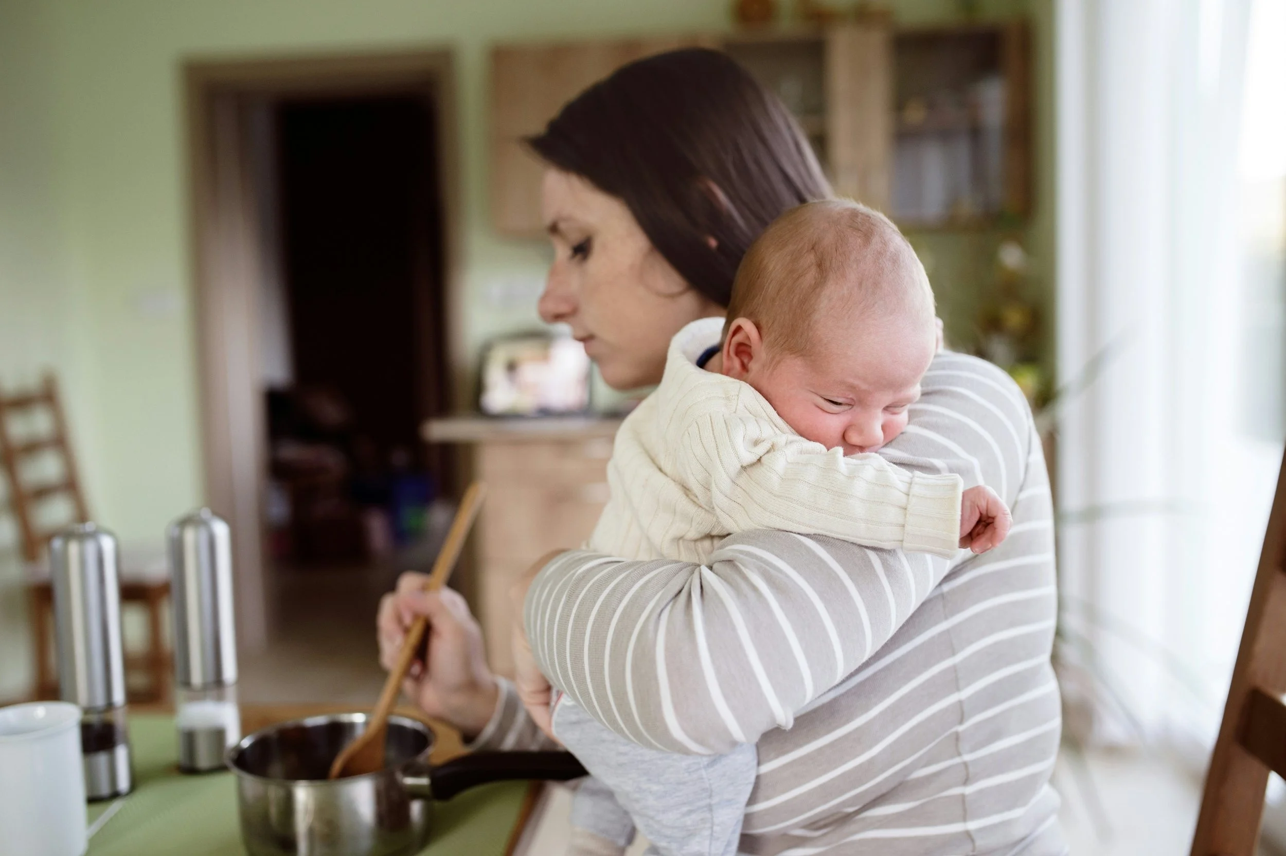 A mother cooking at the stove while holding her infant against her shoulder.