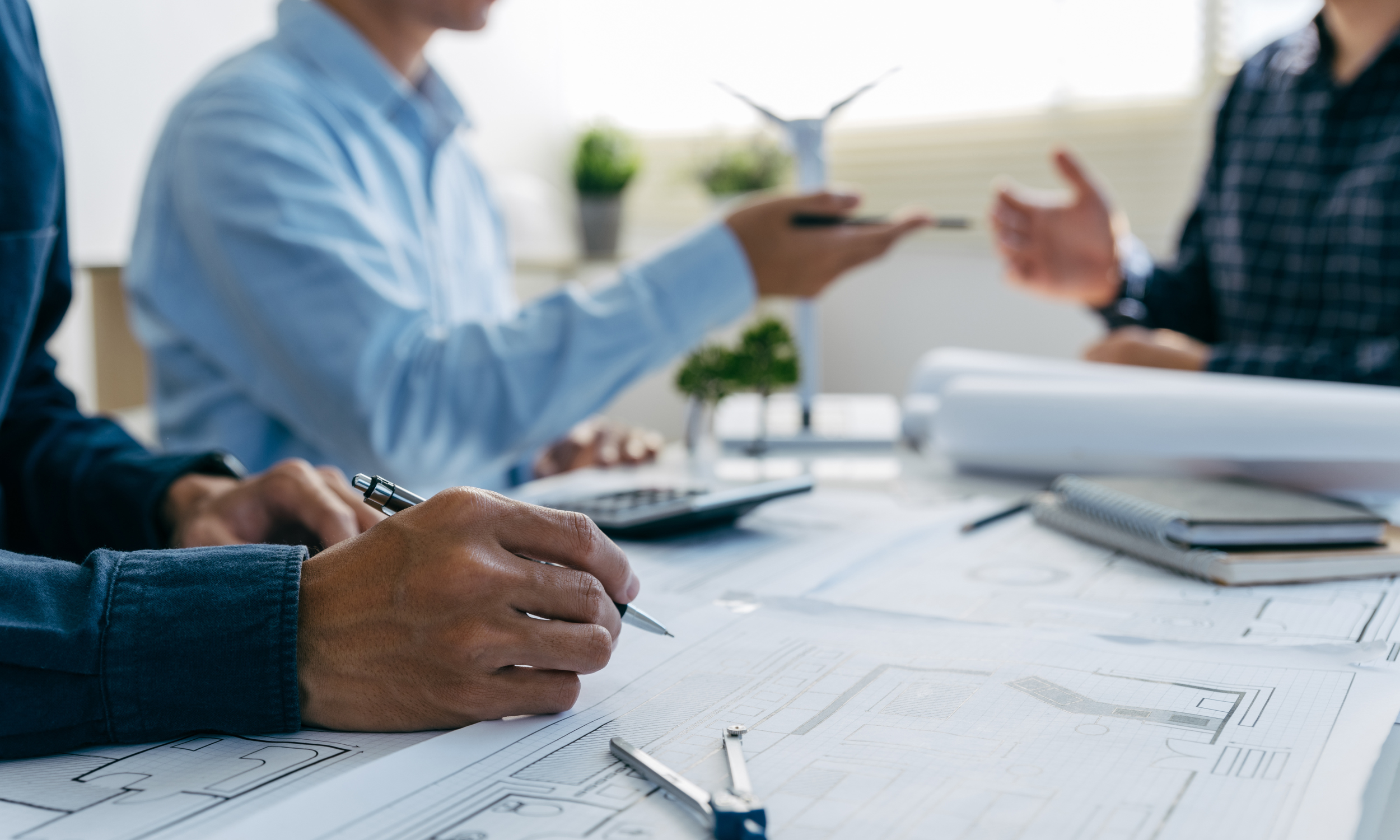 People working on architectural blueprints at a table with office supplies and plants in the background.