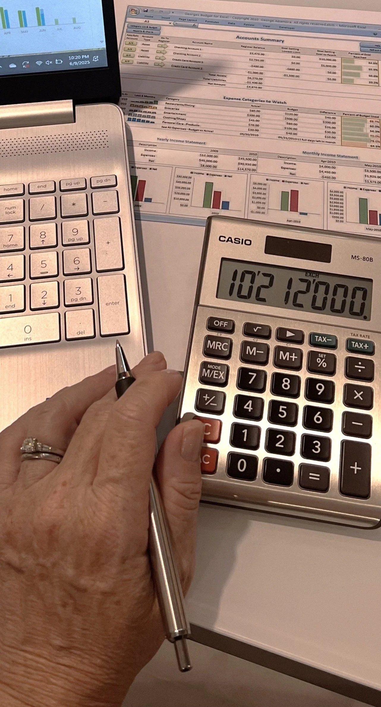 A person's hand holding a calculator showing the number 10212000, with a pen in the hand and laptops and financial documents on the table.