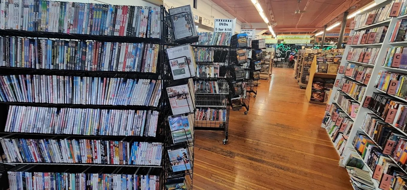 A bookstore aisle with shelves of DVDs on the left, a display rack with miscellaneous items in the middle, and more bookshelves on the right, with a wooden floor and a brightly lit ceiling.