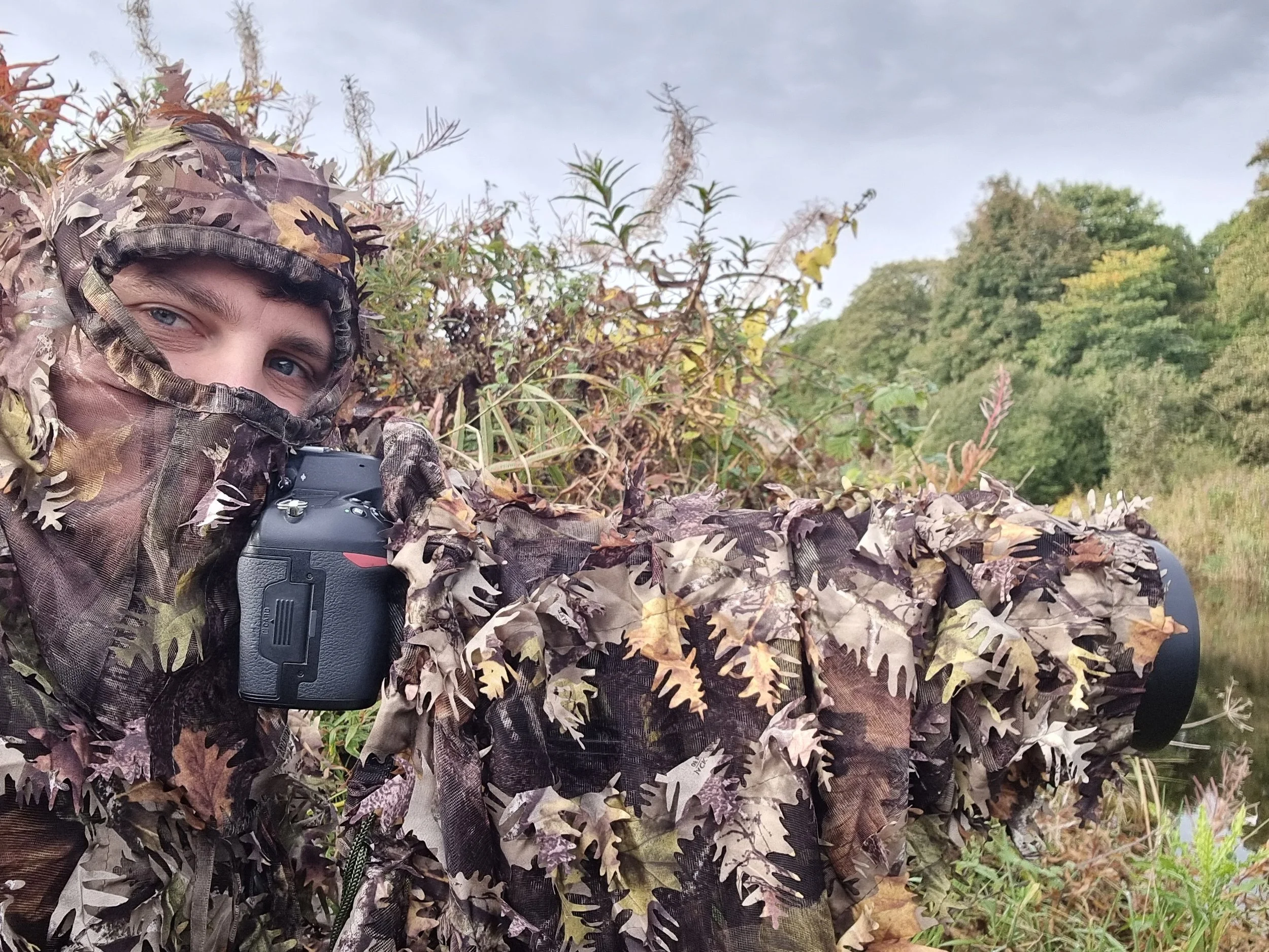 Person in camouflage hunting attire holding a camera, crouching in nature with trees and bushes in the background.
