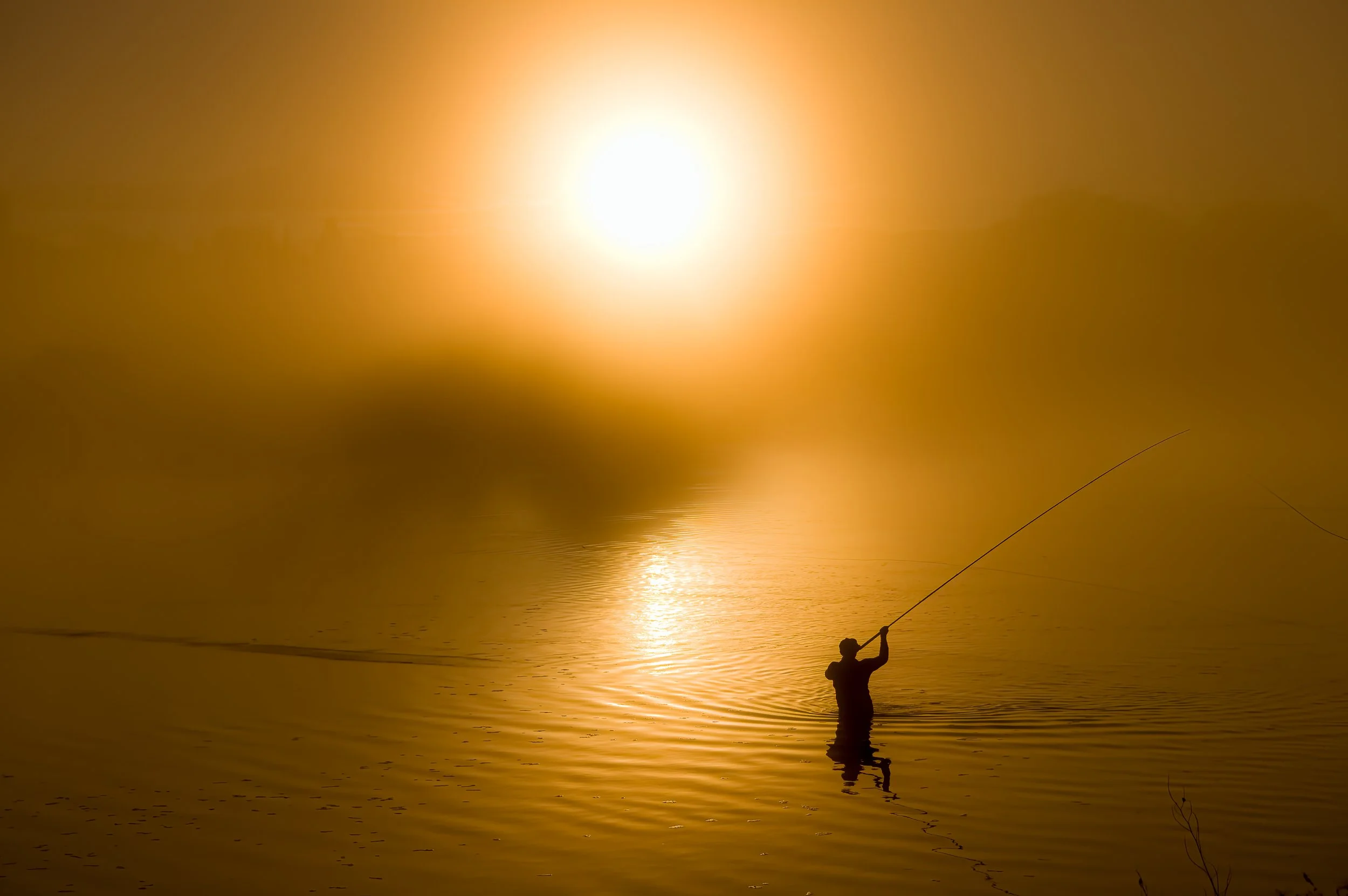 A person fishing in a body of water during sunset or sunrise, with the sun shining brightly in the sky, creating a golden reflection on the water.
