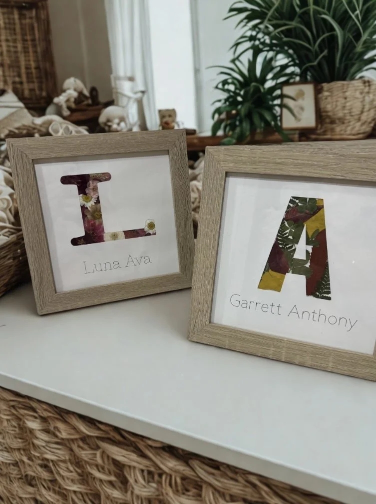 Two frames sit on a table. One has an L cutout with flowers, and the other has an A with leaves. Both have names written under the floral designs