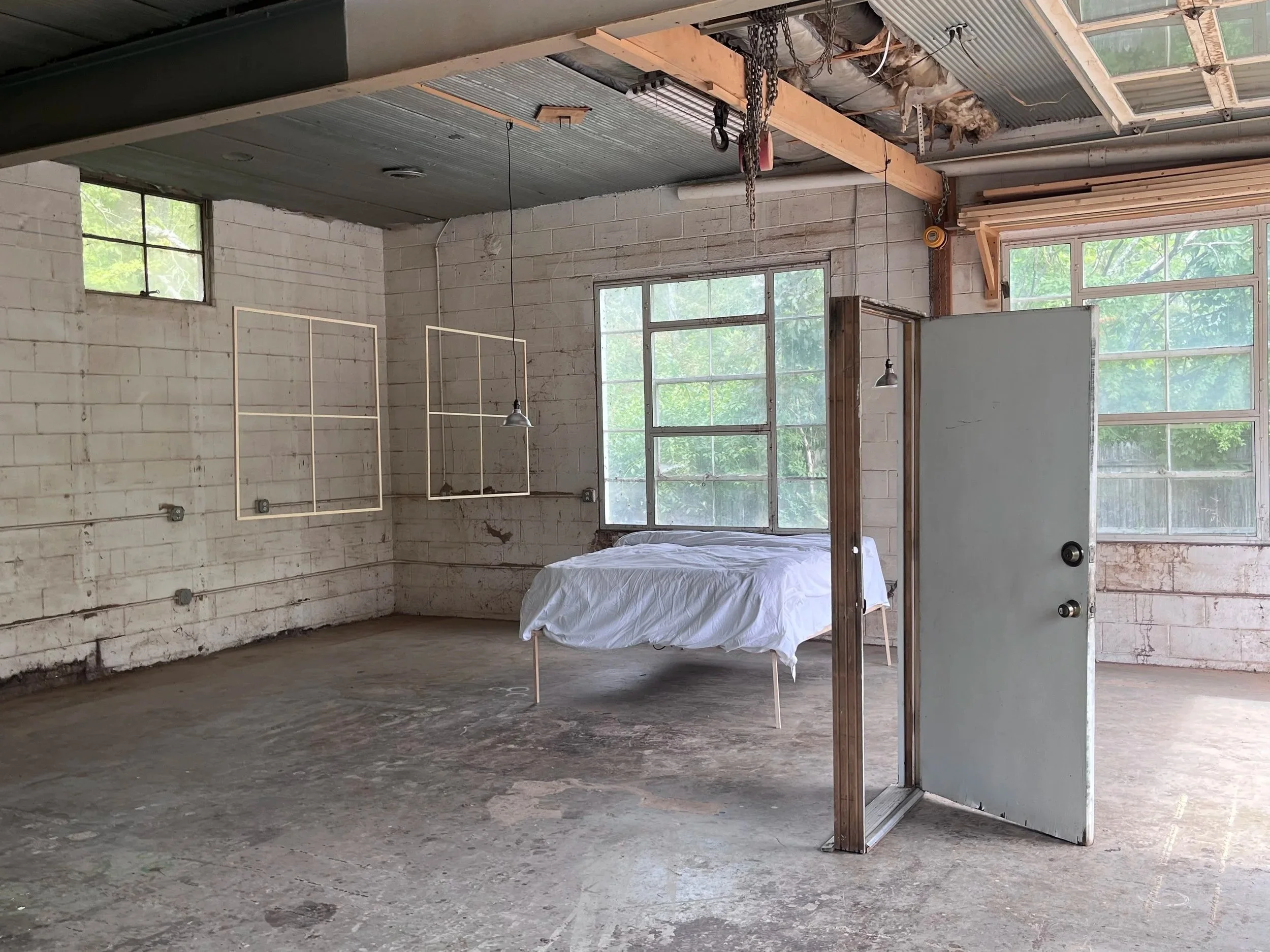 A mostly empty, unfinished room with exposed brick walls, large windows, and a concrete floor. There is a bed with white sheets and a metal frame, and a shower door standing without a shower. Construction materials like chains, wood, and insulation a