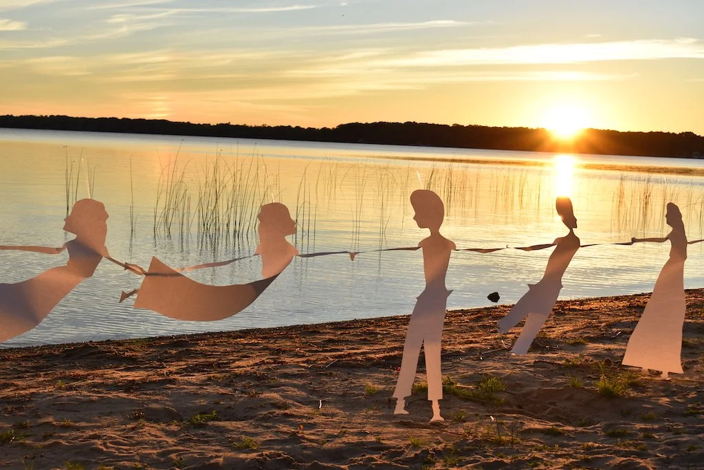 Paper cutout figures of people holding hands along a beach at sunset.