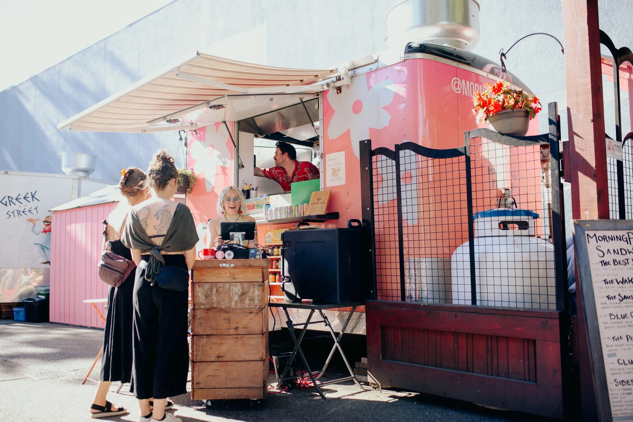 A pink food truck selling sandwiches with customers ordering at the window. The truck has a menu board and potted plants, with a man serving customers and a woman smiling at the counter.