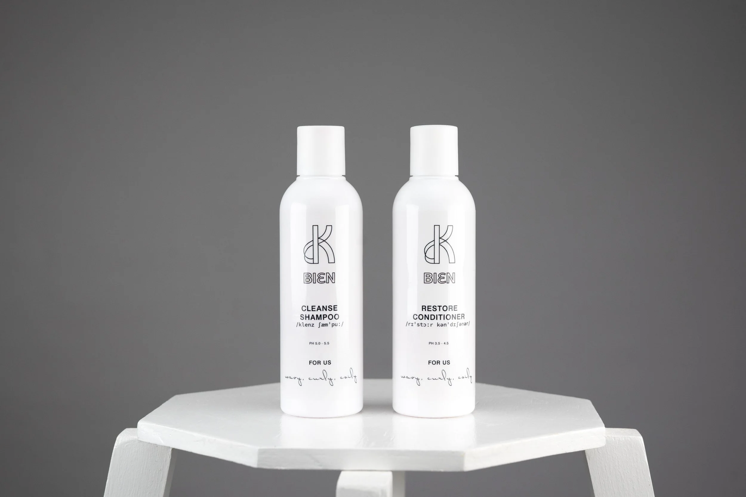 Two white bottles of hair care products, labeled 'K-BIEN' on the front, placed on a white table against a gray background. The bottles are labeled as 'CLEANSE SHAMPOO' and 'RESTORE CONDITIONER'.