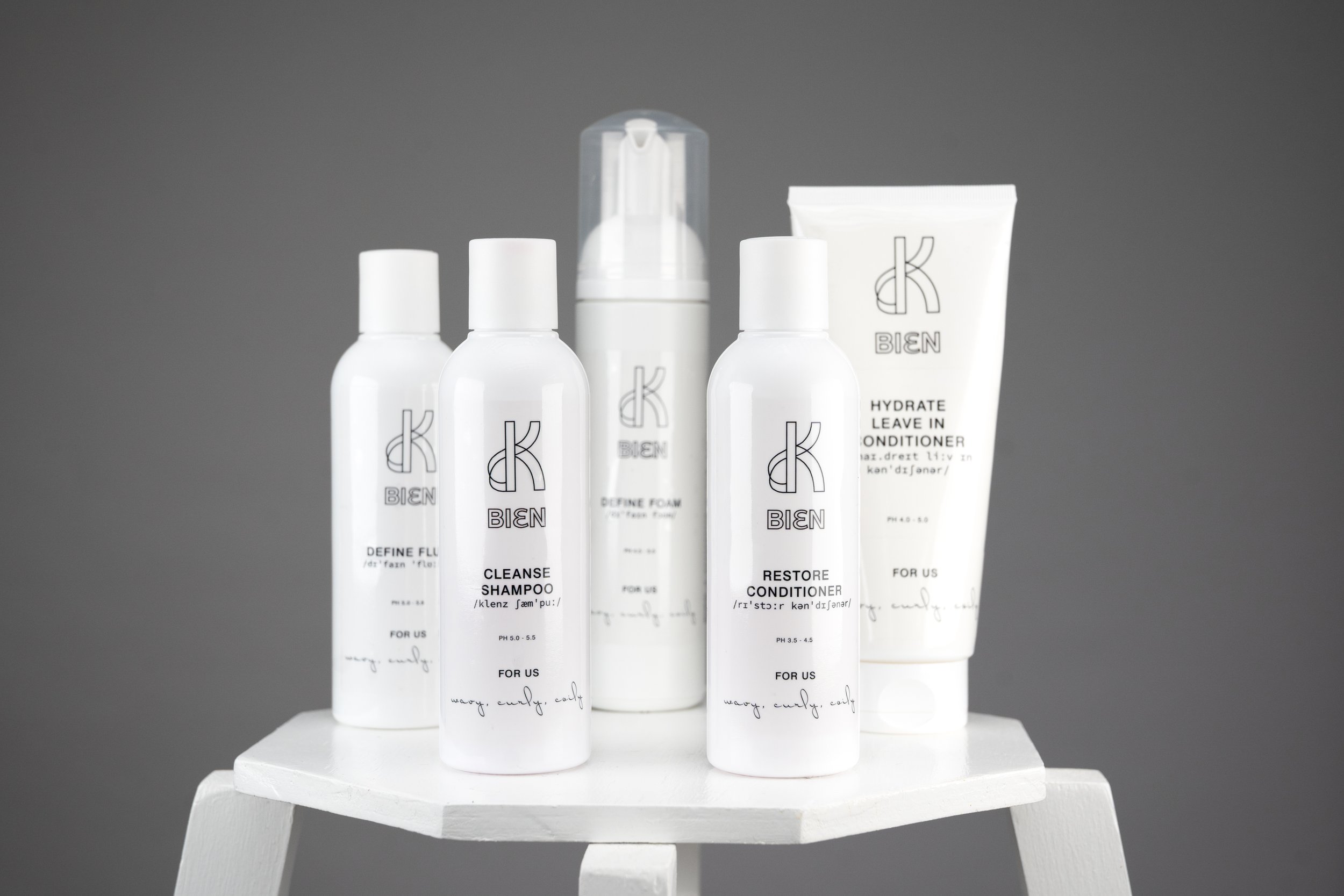 Two white bottles of hair care products, labeled 'K-BIEN' on the front, placed on a white table against a gray background. The bottles are labeled as 'CLEANSE SHAMPOO' and 'RESTORE CONDITIONER'.