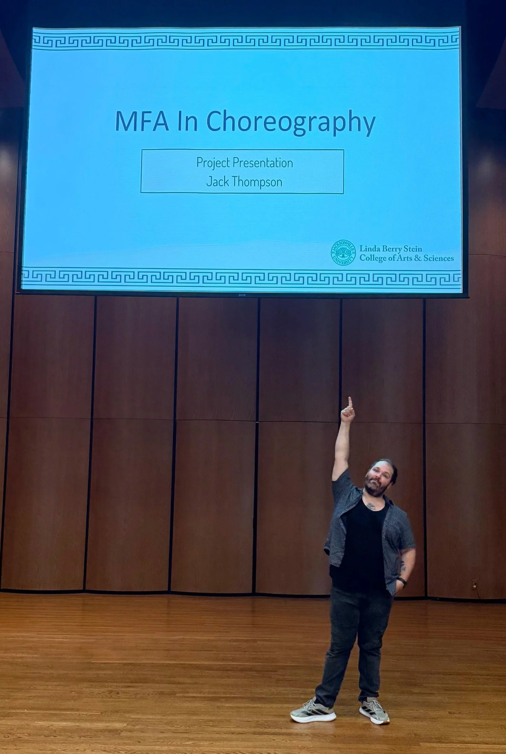 A man with a beard and tattoos on his arm is standing in a wooden-floored room, pointing up at a large screen. The screen displays a presentation slide titled 'MFA In Choreography' by Jack Thompson, with the logo of the University of Jacksonville and the text 'Linda Berry Stein College of Arts & Sciences' at the bottom right corner.
