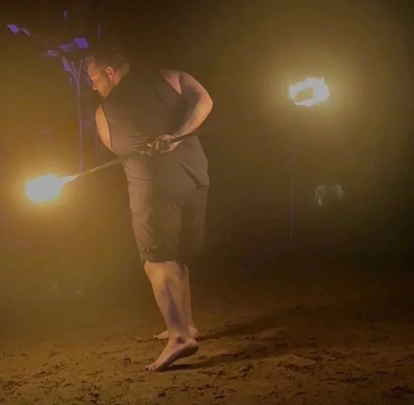 A man practicing fire spinning with a flaming torch in a dark environment, standing barefoot on a dirt floor.