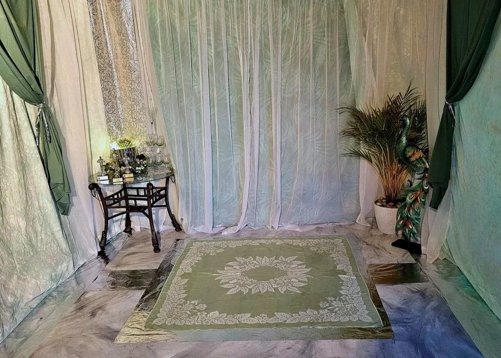 A small vintage-style table with glassware and decorative items, a green patterned area rug, potted plant, and curtains in shades of green and cream in a room with marble flooring.
