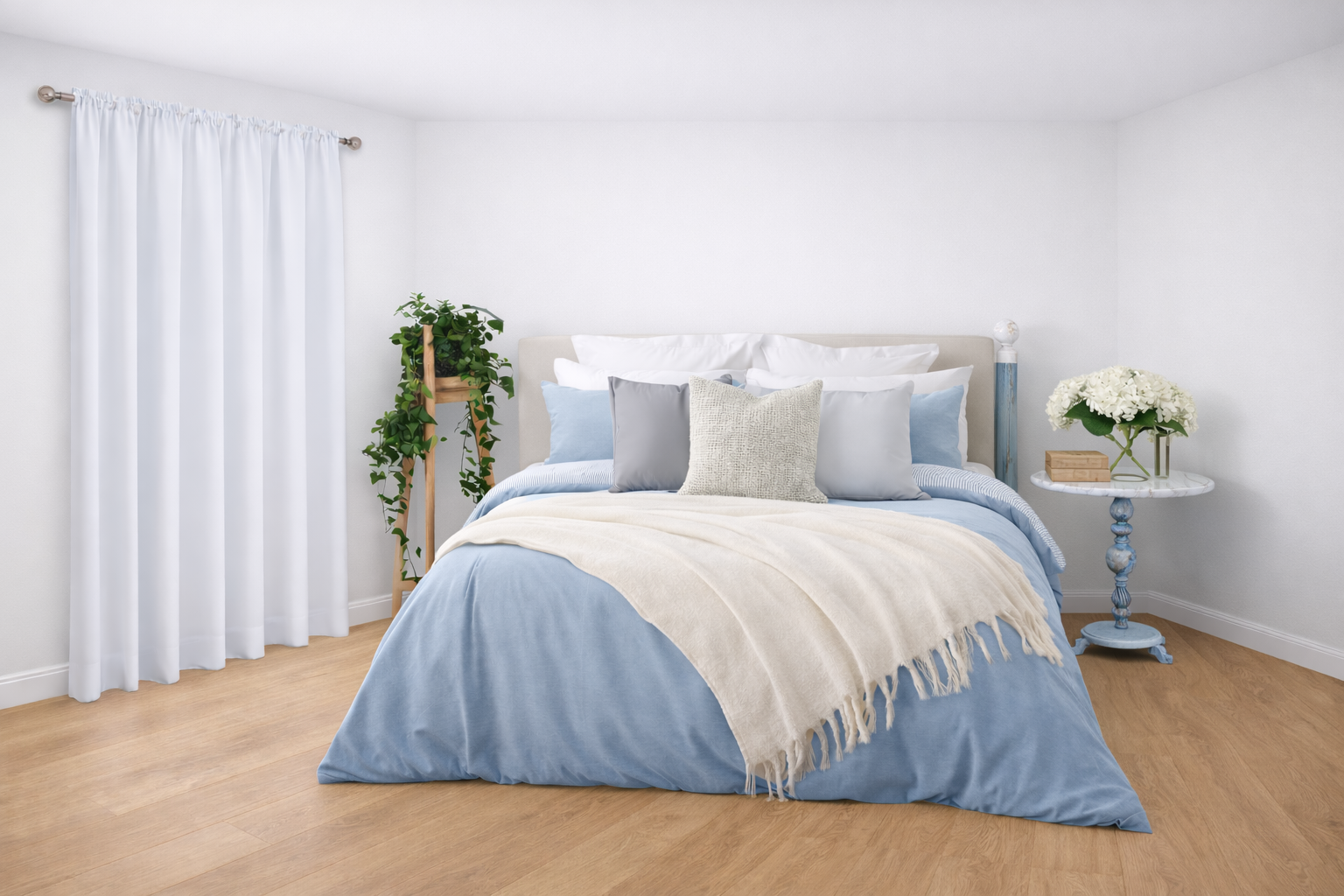 A neatly made bed with light blue bedding, multiple pillows including white, gray, and textured beige pillows, a cream throw blanket, a small wooden side table with a bouquet of white flowers and a small wooden box, a potted plant on a wooden stand, white curtains, and light wood flooring.