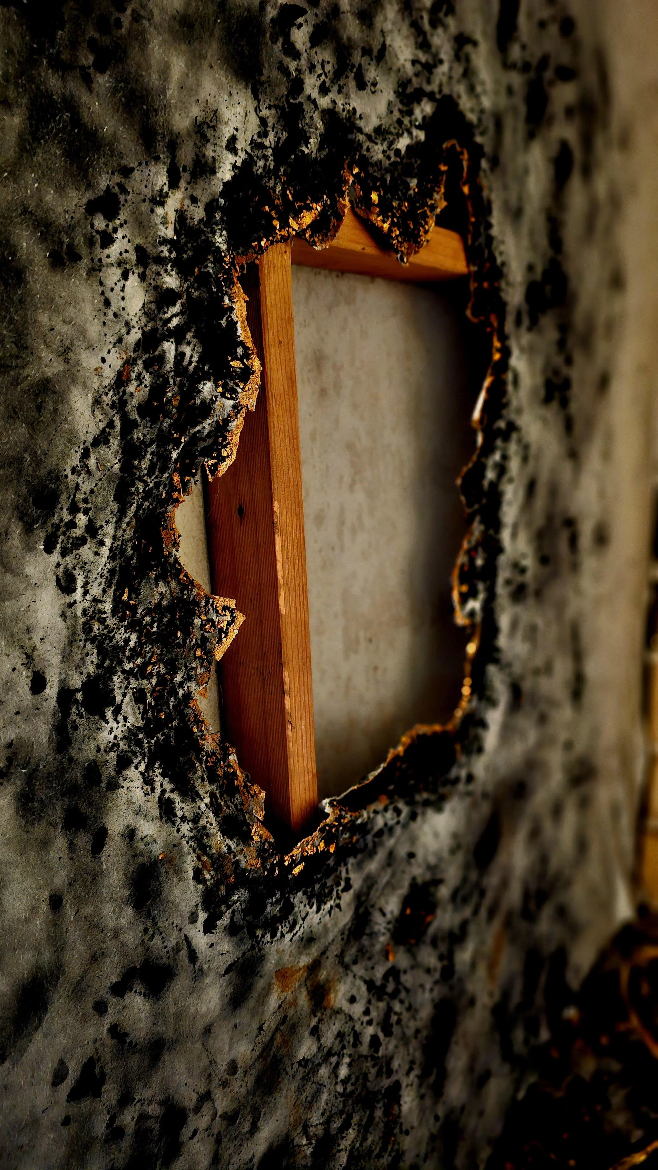 A burnt and charred wall with a large hole in the center, revealing a wooden beam and beige wall behind it.