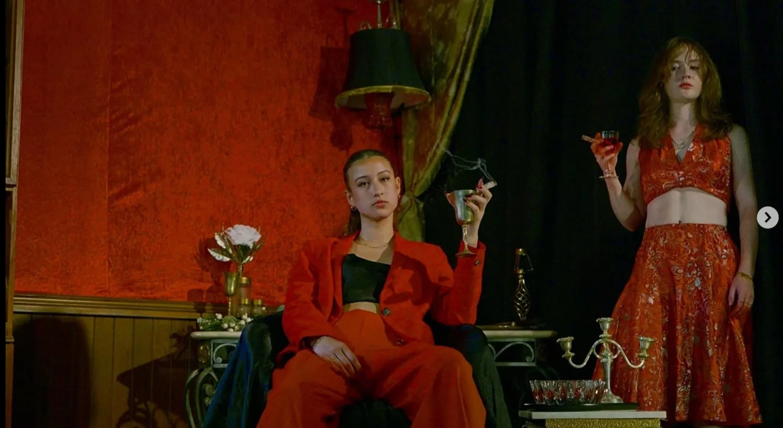 Two women in stylish red outfits in a dimly lit room. One is seated in a chair holding a drink, while the other stands holding a glass of red wine. The room has a vintage vibe with dark curtains, a green lampshade, and elegant decor.