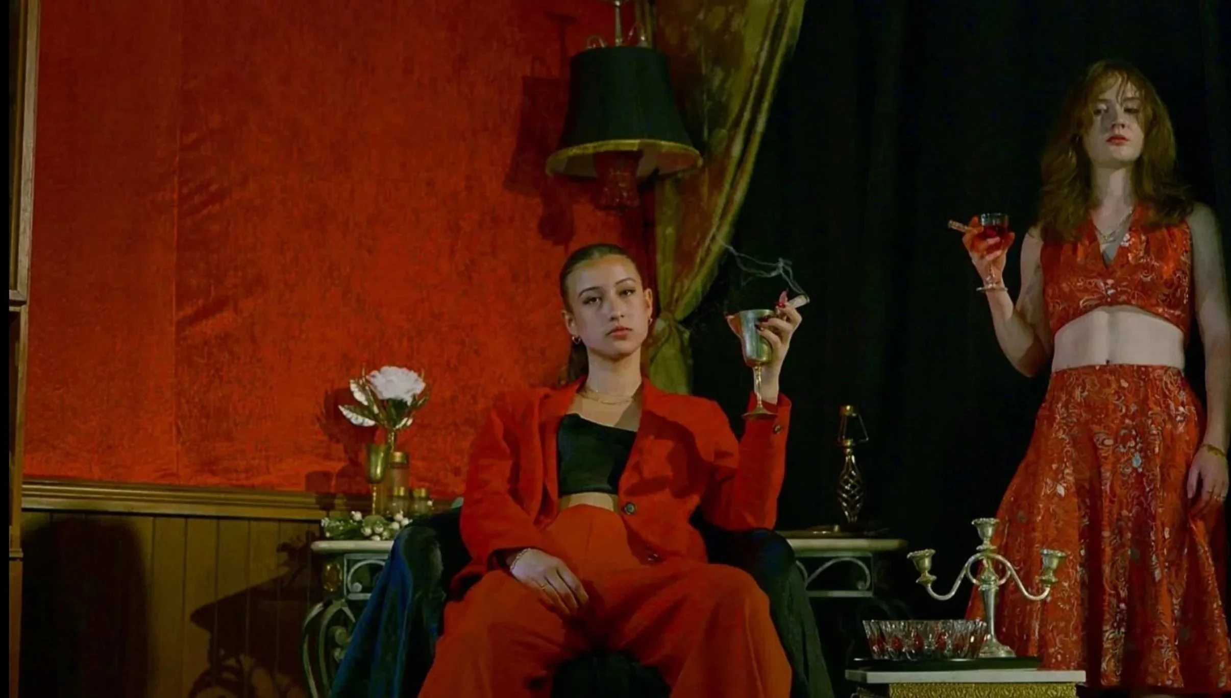 Two women in red outfits in a dimly lit room with vintage decor. One woman is sitting on a black chair holding a lit cigarette and a drink, while the other stands holding a glass. There is a dark drape behind them and a side table with a floral arrangement and decorative items.