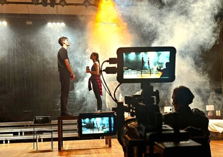 Two performers on stage, one man and one woman, are being filmed by a camera operator during a rehearsal or shoot in a theater or studio with stage smoke and lighting.