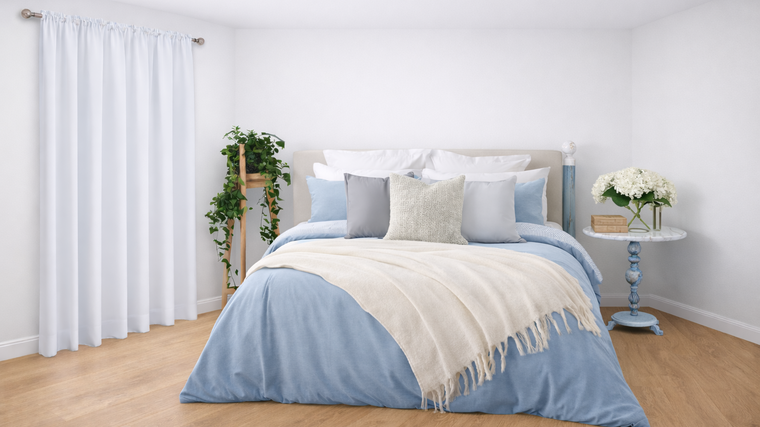 A neatly made bed with blue and white bedding, multiple pillows, and a cream throw blanket in a bright bedroom with wooden floors, white walls, a white curtain, a potted plant on a wooden stand, and a small blue nightstand with white flowers and books.