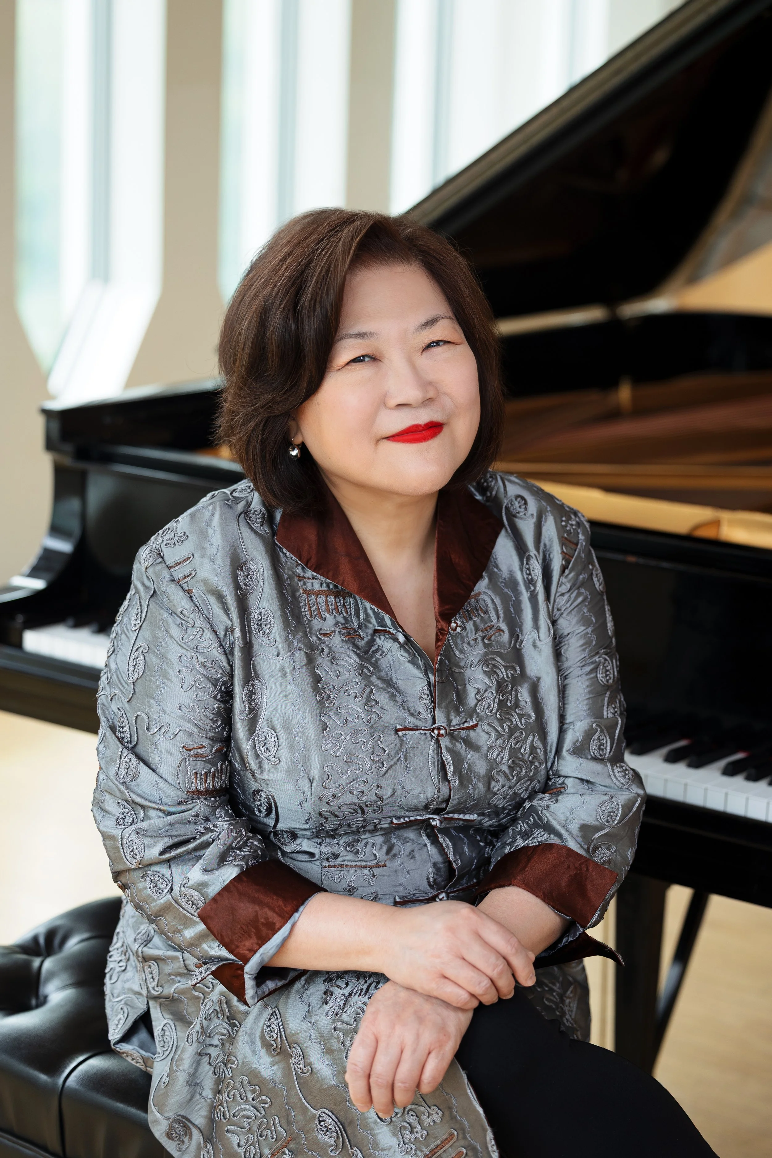 A woman with short dark hair wearing a silver and brown traditional Asian silk blouse, sitting on a black bench in front of a grand piano in a bright room with large windows.