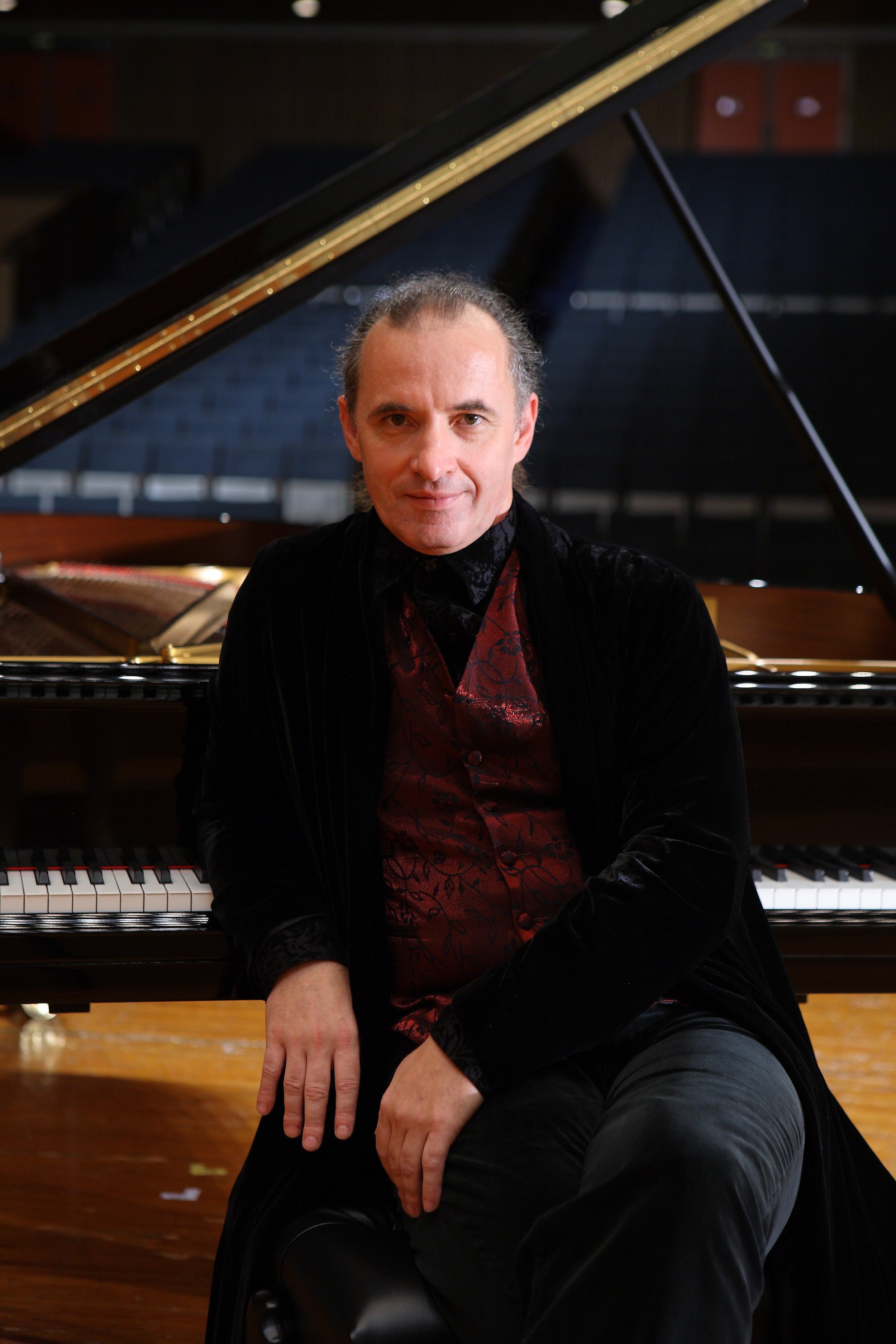 A man sitting in front of a grand piano, wearing a dark velvet jacket and red patterned vest, in a dimly lit music studio or concert hall.