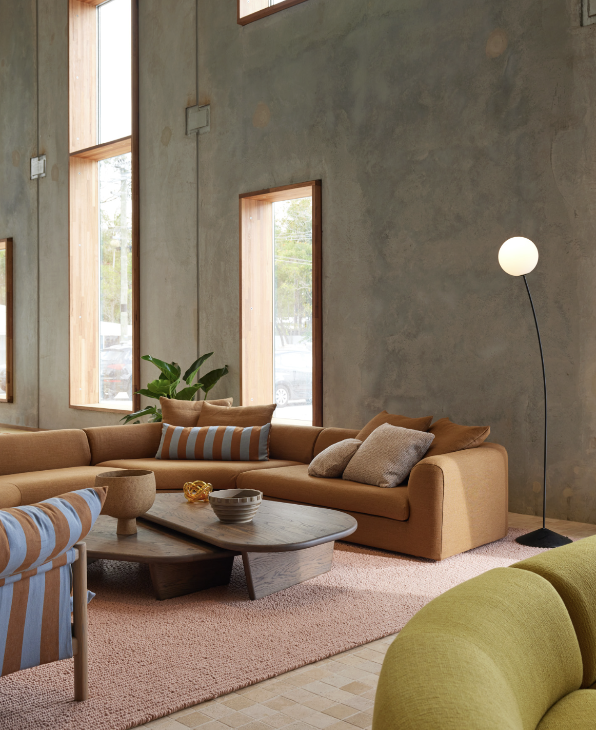 A modern living room with a large mustard-colored sofa, a low wooden coffee table, and multiple patterned pillows. There is a tall, white floor lamp, a pink textured rug, and large windows with wooden trim that let in natural light. A green plant is near the windows and the walls are textured with a concrete finish.