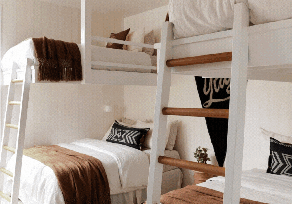 Two sets of bunk beds with white frames, beige and black pillows, brown blankets, and a small plant in a pot on a side table.