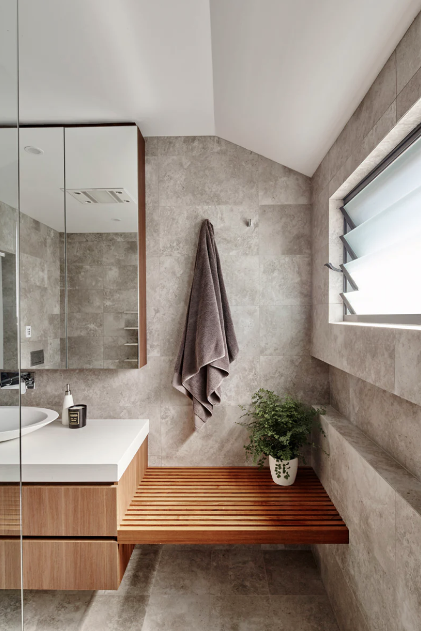 Modern bathroom with beige tiled walls, a wooden bench, potted plant, gray towel hanging, and a window with louvered glass panes.