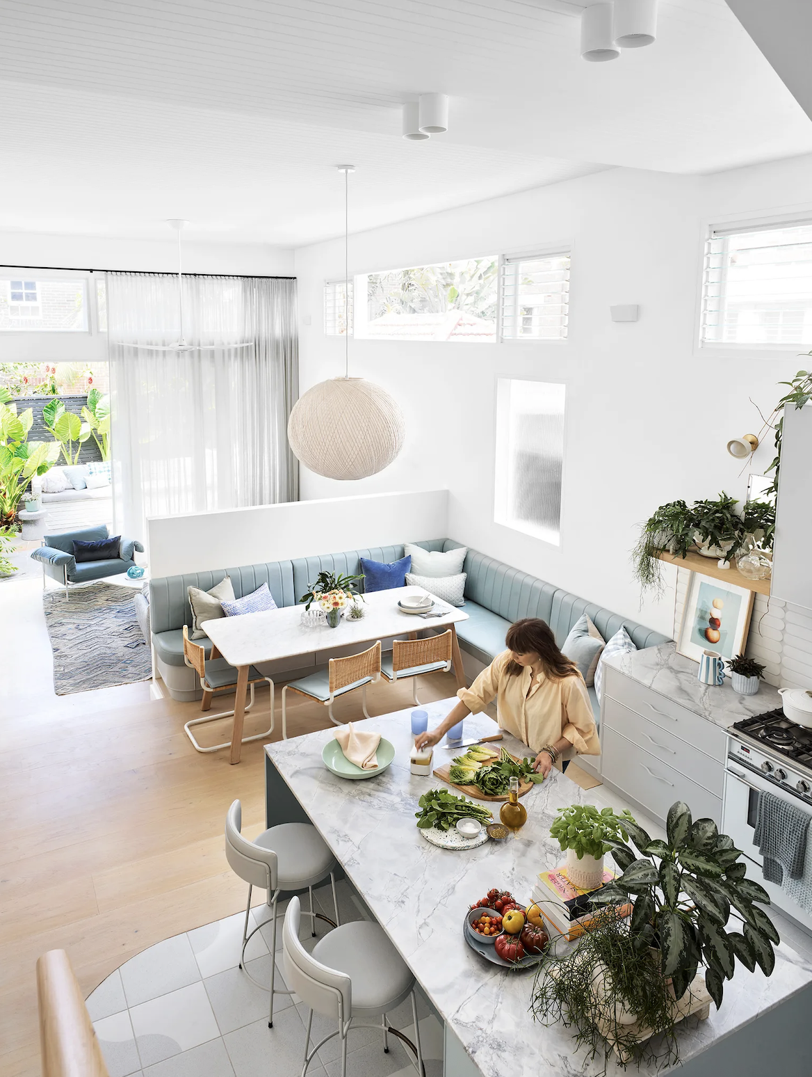 Bright, modern open-plan kitchen and dining area with white walls and ceiling, a marble kitchen island, a woman preparing vegetables, and a cozy living space with a cushioned dining nook and outdoor patio.