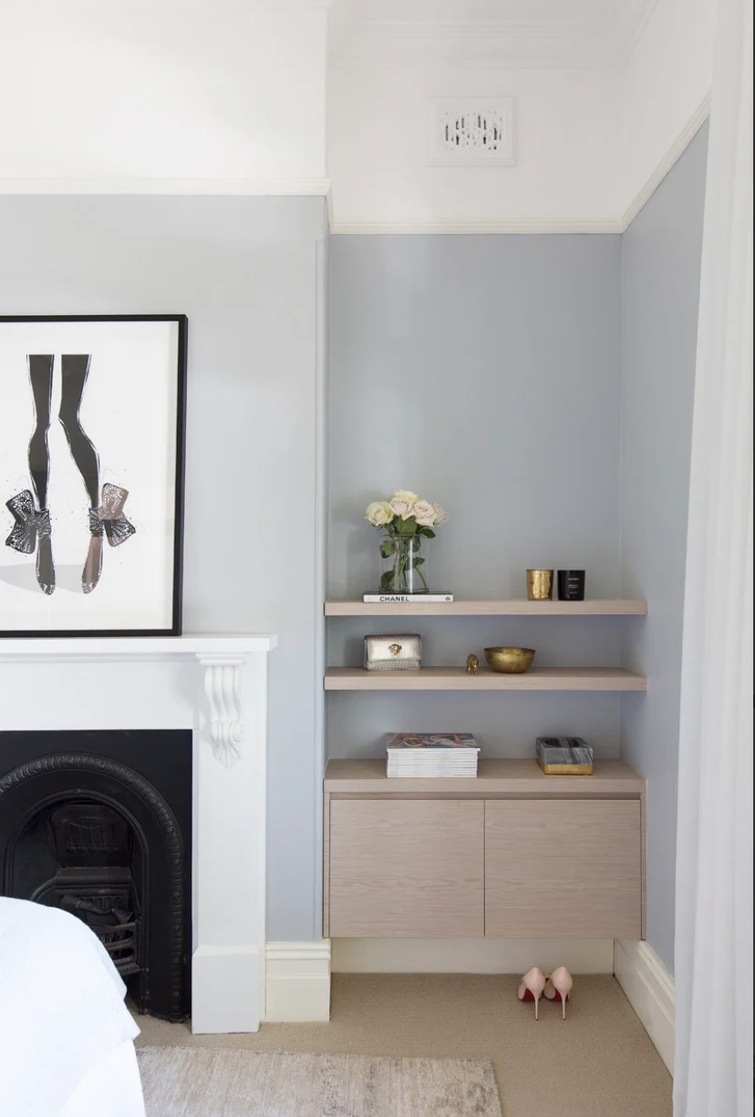 Corner of a room with light blue walls, a built-in white fireplace with decorative molding, and a wooden shelf with decorative items including a vase of flowers, candles, and jewelry boxes. Pink high heel shoes are on the carpeted floor near the shelf.