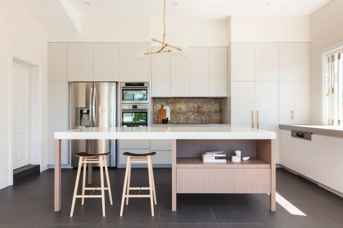 Modern white kitchen with a large island, two stools, stainless steel appliances, and gold accents.