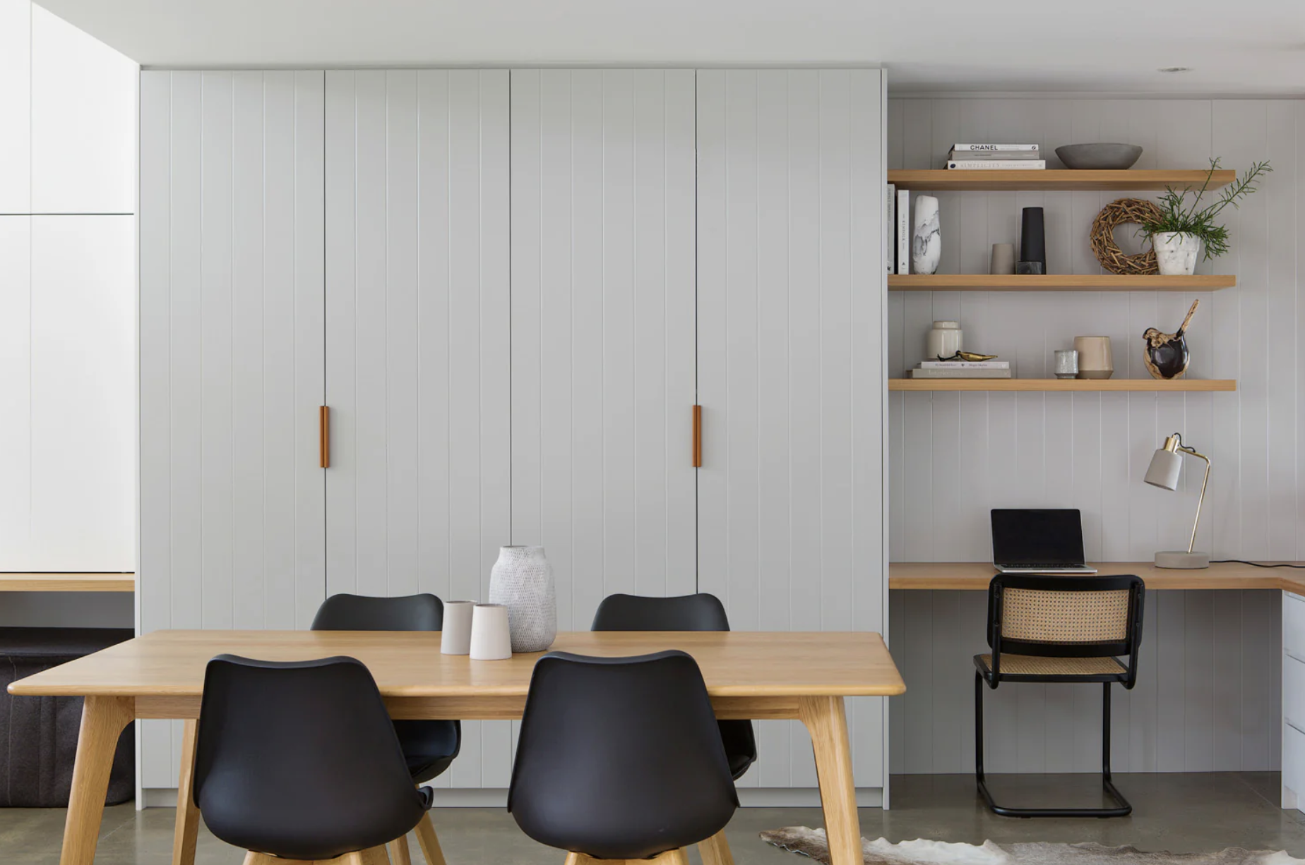 Modern dining and workspace with a wooden table, black chairs, white wall with peninsula, and open floating shelves with books, vases, and decorative items.