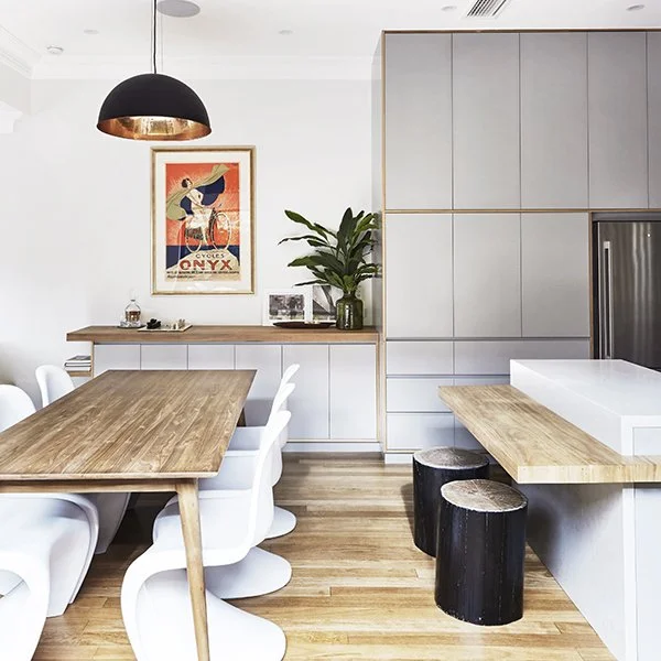 Modern kitchen and dining area with a wooden table and white chairs, a black pendant light, and cabinetry with a built-in fridge. There is a framed vintage poster, a potted plant, and black stools nearby.