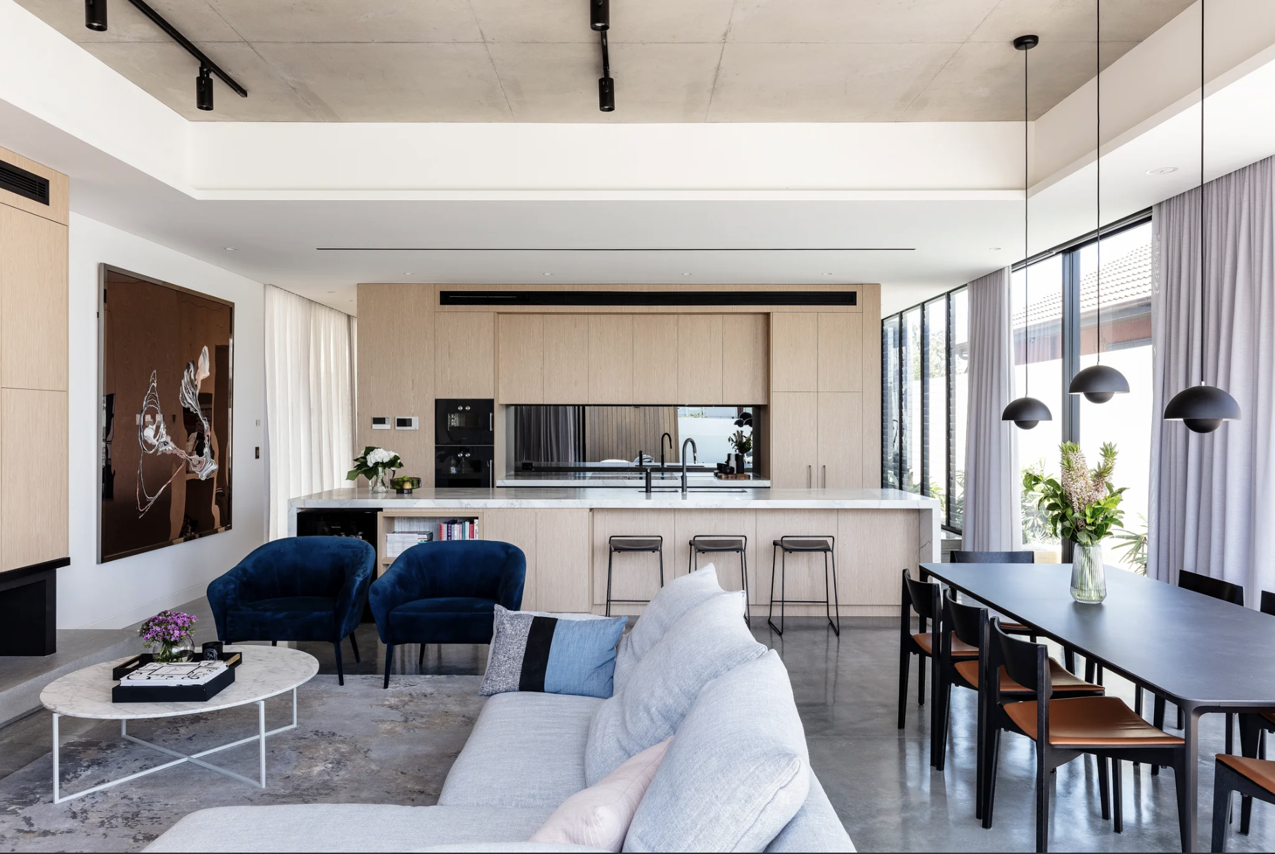 Modern open-plan living room and kitchen with a dining table, blue and gray furniture, large windows with curtains, and minimalist decor.