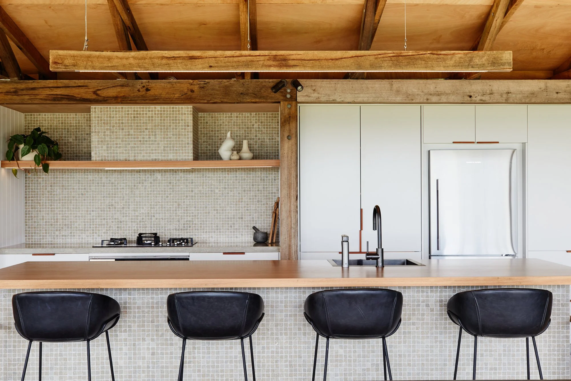 Modern kitchen with a wooden ceiling, black chairs at a wooden countertop, white cabinets, and a mosaic tile backsplash.