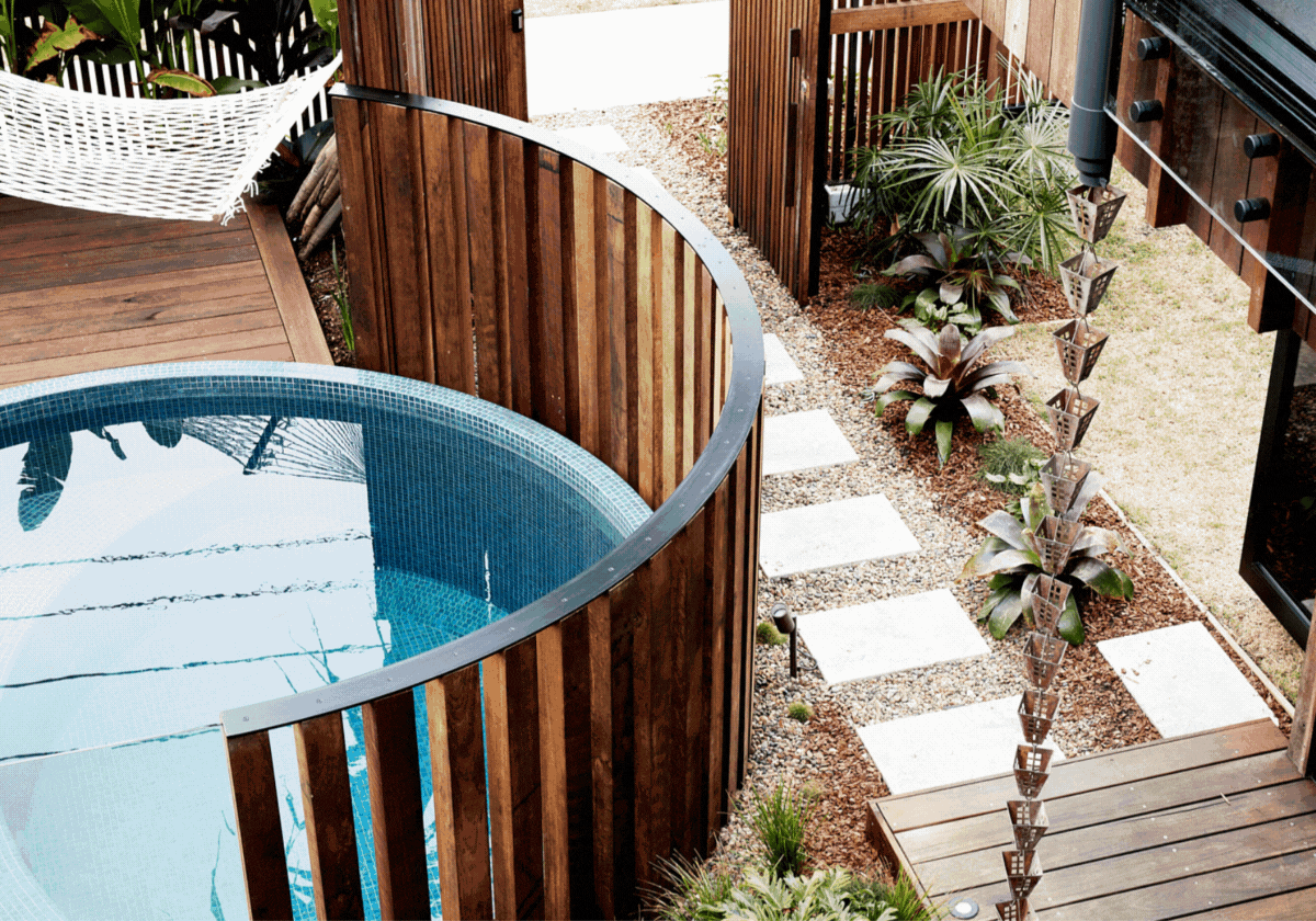 A view of a balcony with a hot tub surrounded by a wooden fence, stepping stones leading to a garden with plants, and part of a black outdoor grill.