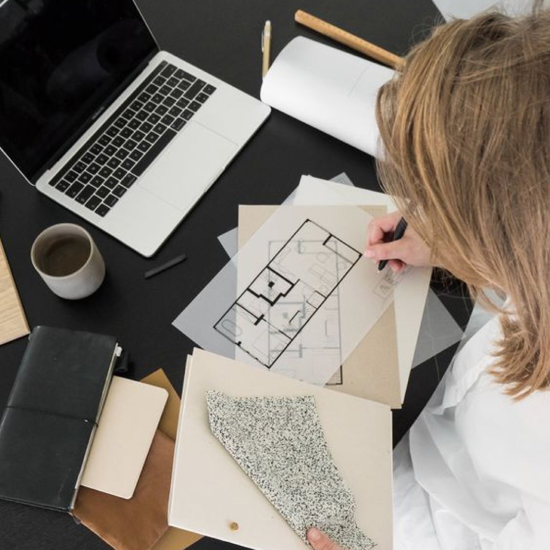 A person working at a desk with architectural plans, a laptop, a notebook, a coffee mug, and color samples.