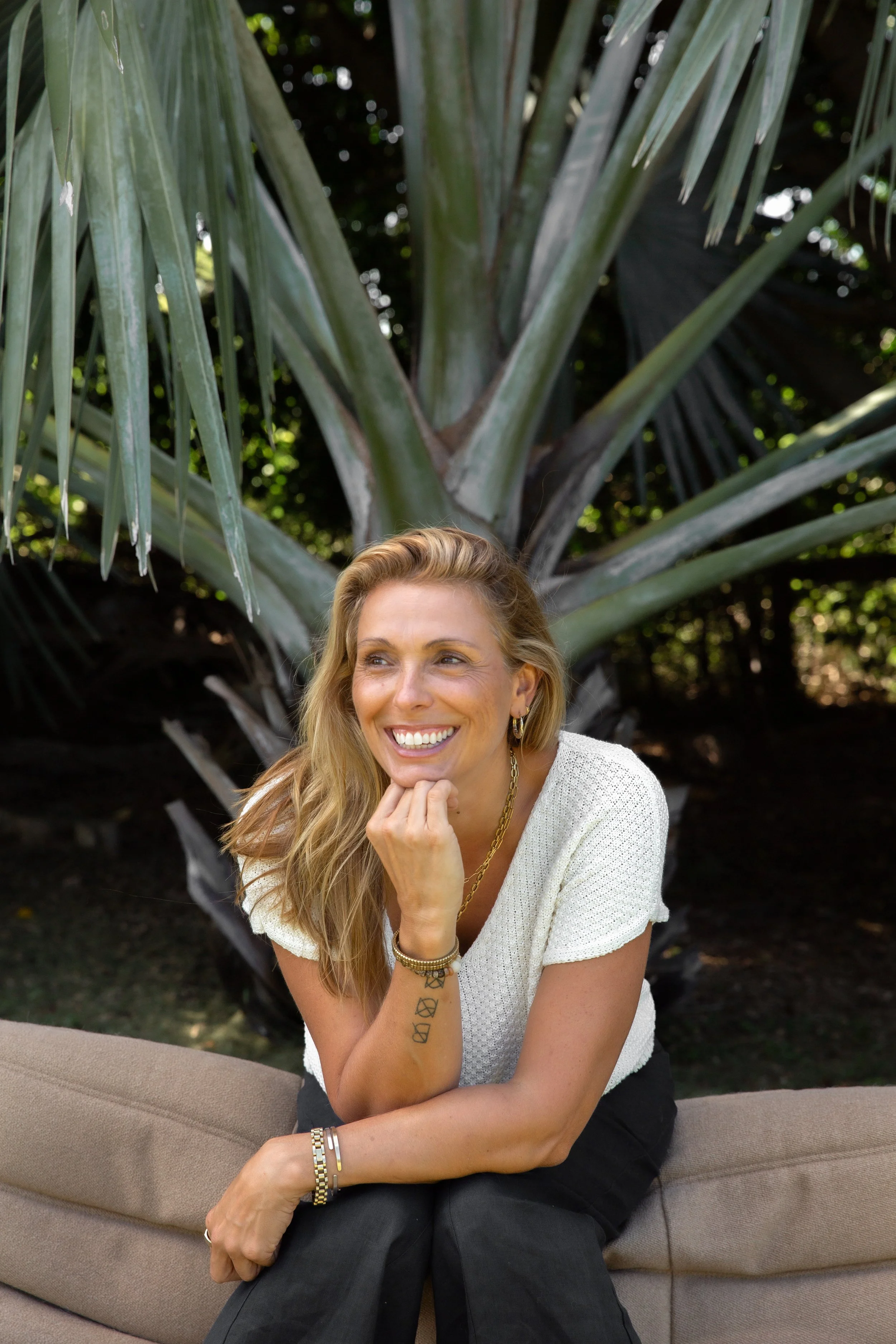 A woman sitting outdoors smiling with her chin resting on her hand, in front of a large palm tree.