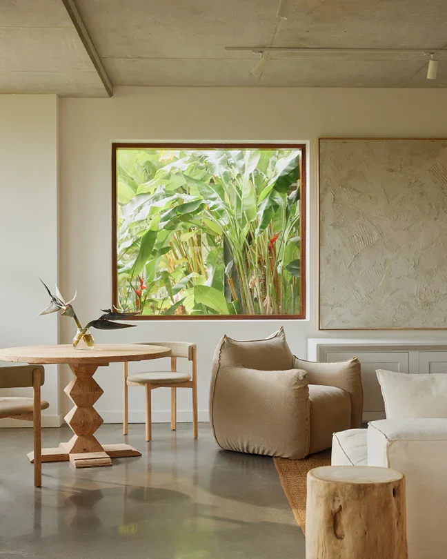 Interior of a modern living room with natural light, featuring a wooden table, beige armchair, white sofa, and a large window showing lush green tropical plants outside.