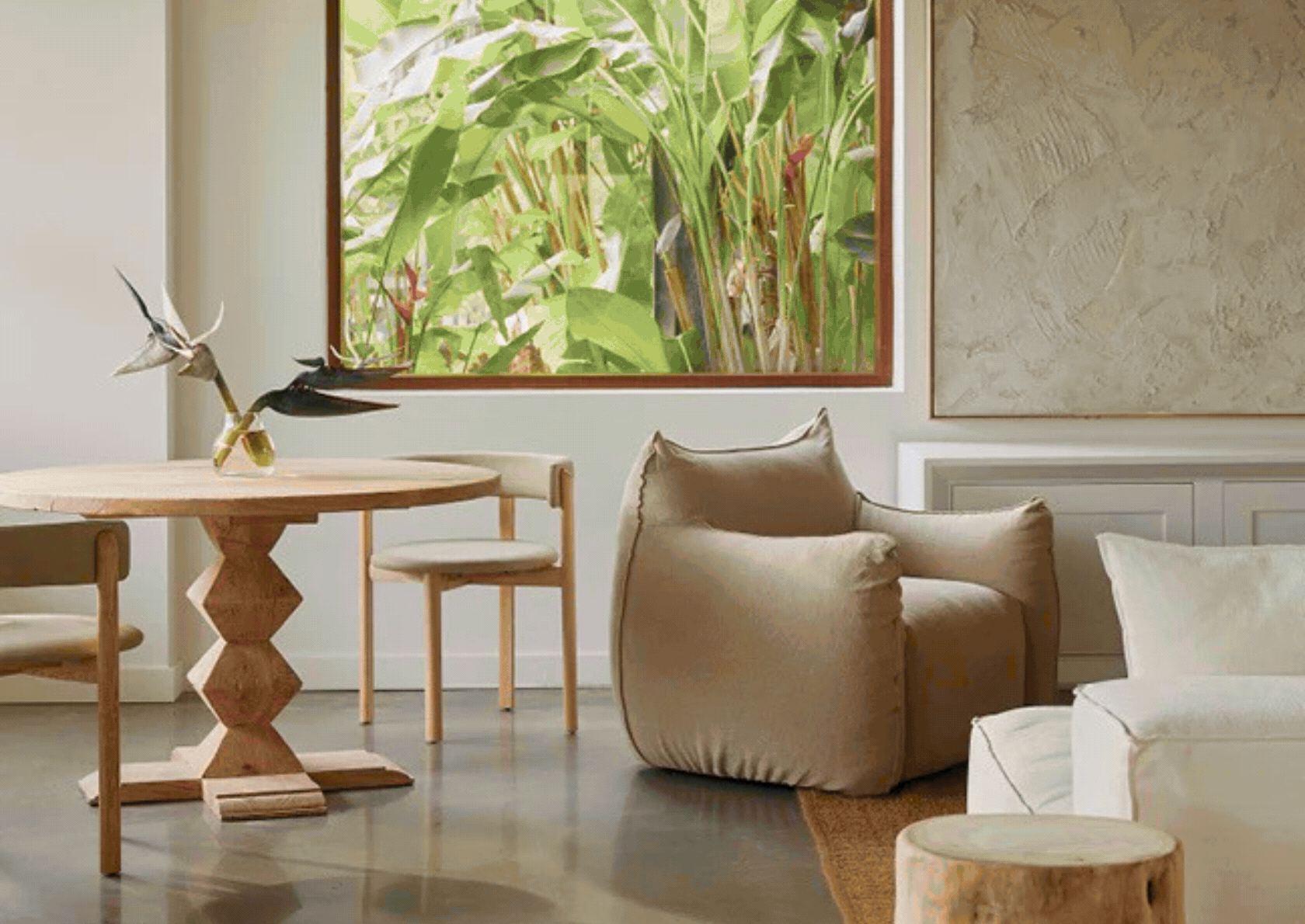 Living room with beige armchair, white sofa, wooden table with decorative bird sculpture, and large window with green plants outside.