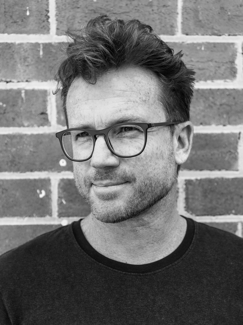 Black and white portrait of a man with glasses, short tousled hair, and a slight smile, standing against a brick wall.