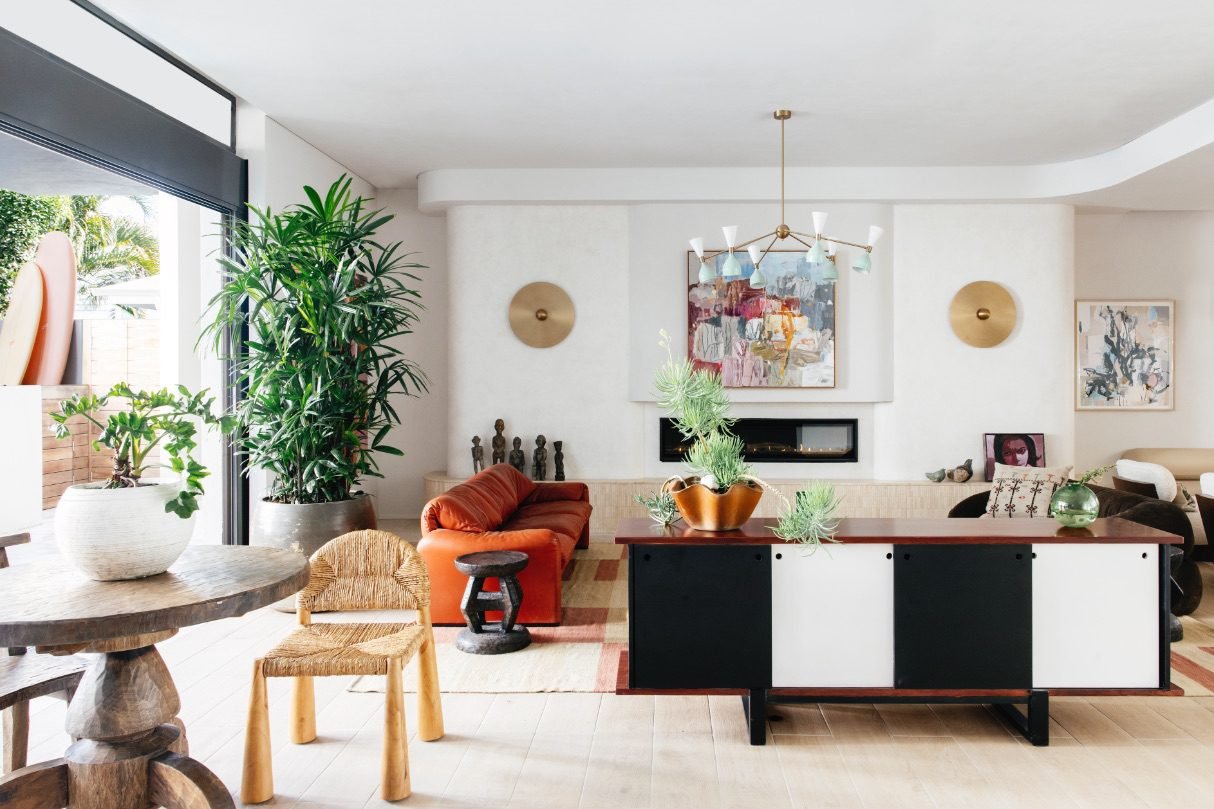 Living room with an abstract painting above a fireplace, a modern chandelier, a red sofa, a large green plant, two wall sconces, a wooden side table, and a black-and-white sideboard with plants and decorative items.